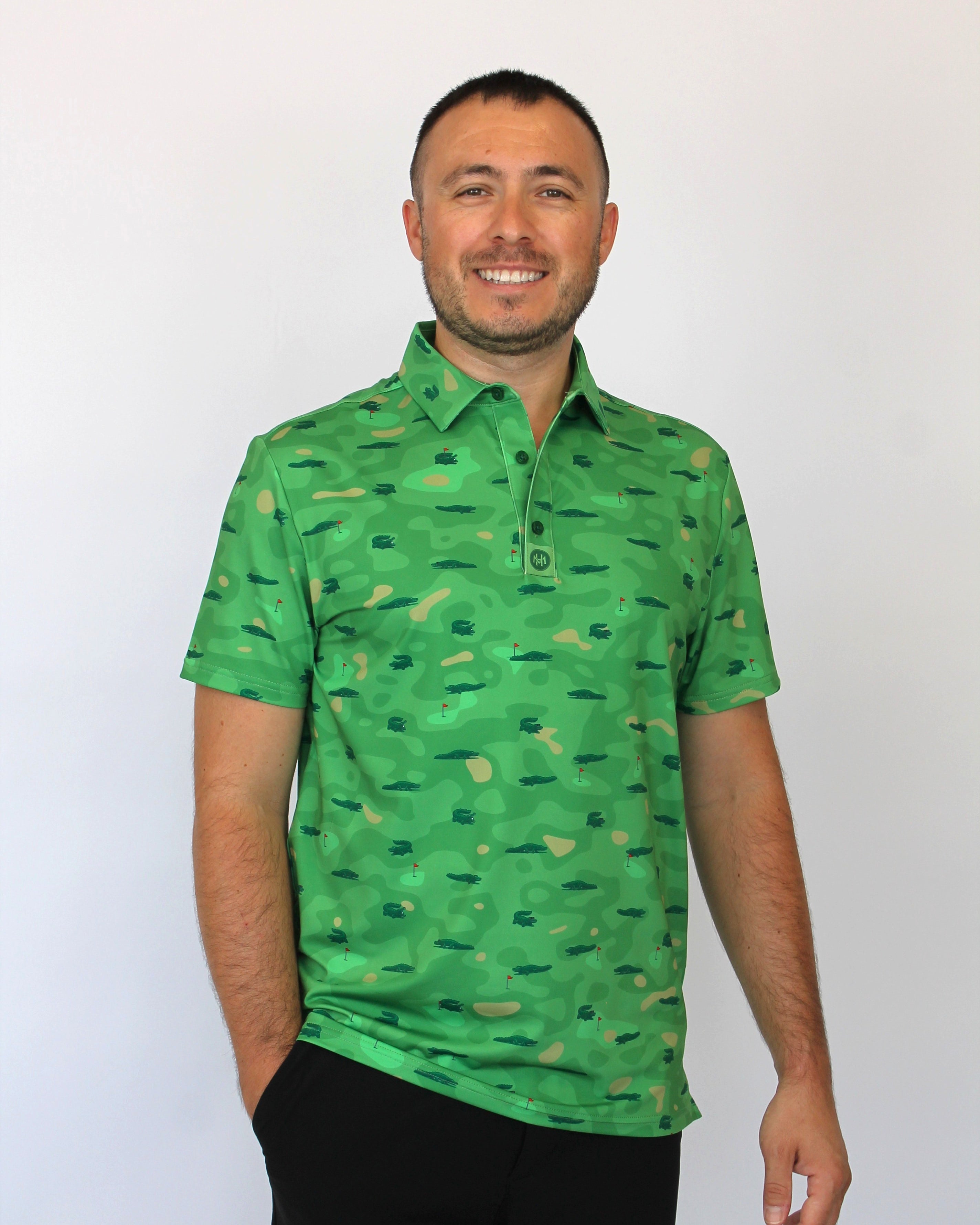 A man wearing a green short-sleeve polo shirt with an all-over camouflage pattern and Alligators on a golf course some with sunglasses, some with a golf ball in their mouth, paired with black pants, standing against a plain white background.