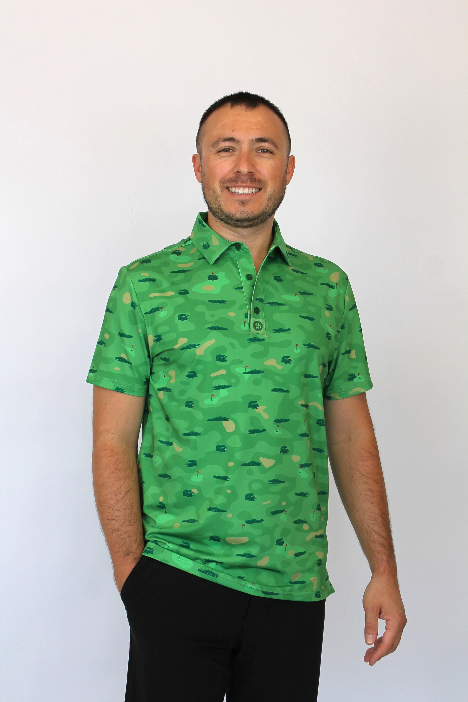 A man wearing a green short-sleeve polo shirt with an all-over camouflage pattern and Alligators on a golf course some with sunglasses, some with a golf ball in their mouth, paired with black pants, standing against a plain white background.