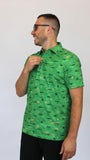 A man wearing a bright camo green short-sleeve button-up shirt with a repeating Alligators on a golf course pattern, paired with black pants and sunglasses, standing against a plain light gray background.