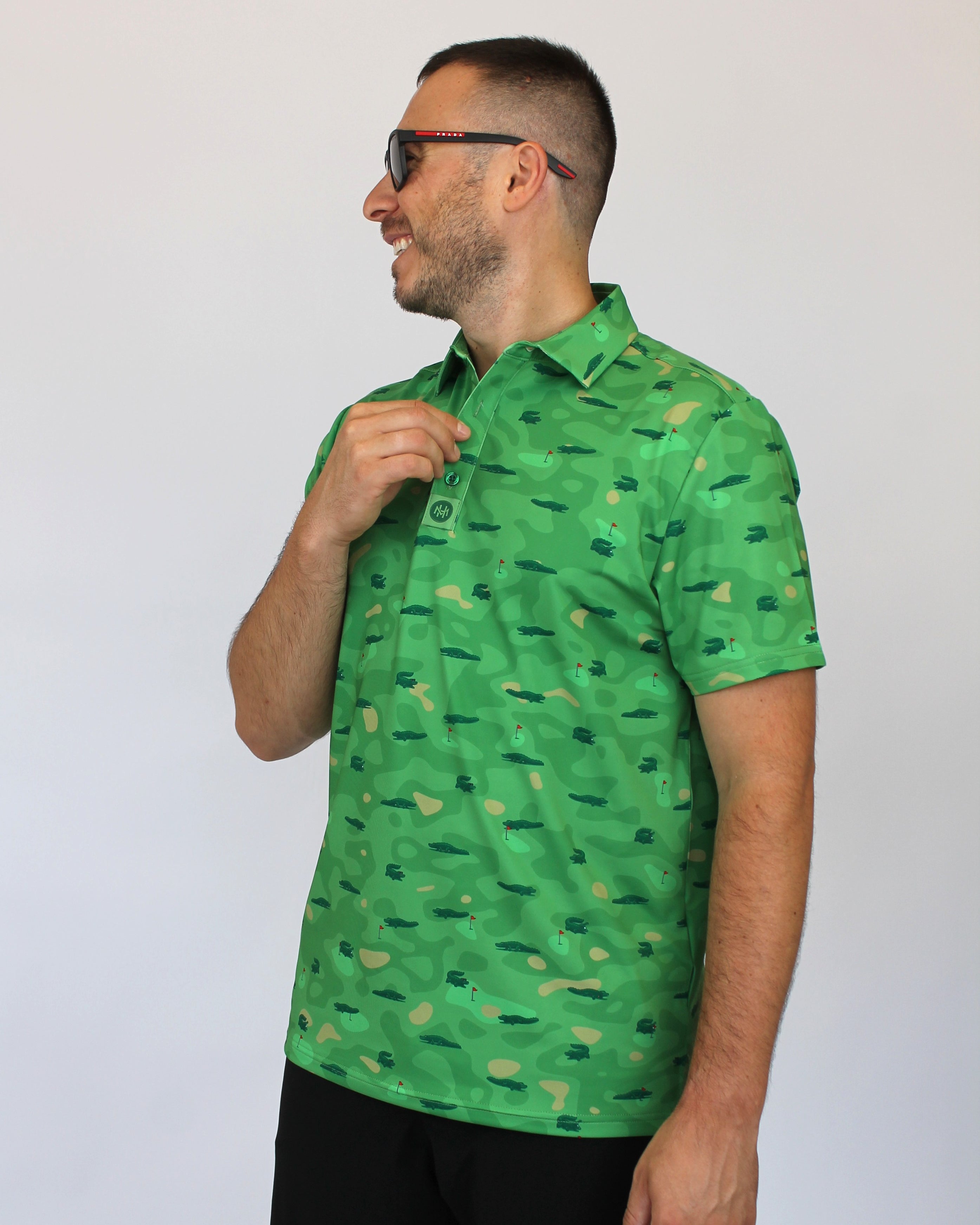 A man wearing a bright camo green short-sleeve button-up shirt with a repeating Alligators on a golf course pattern, paired with black pants and sunglasses, standing against a plain light gray background.