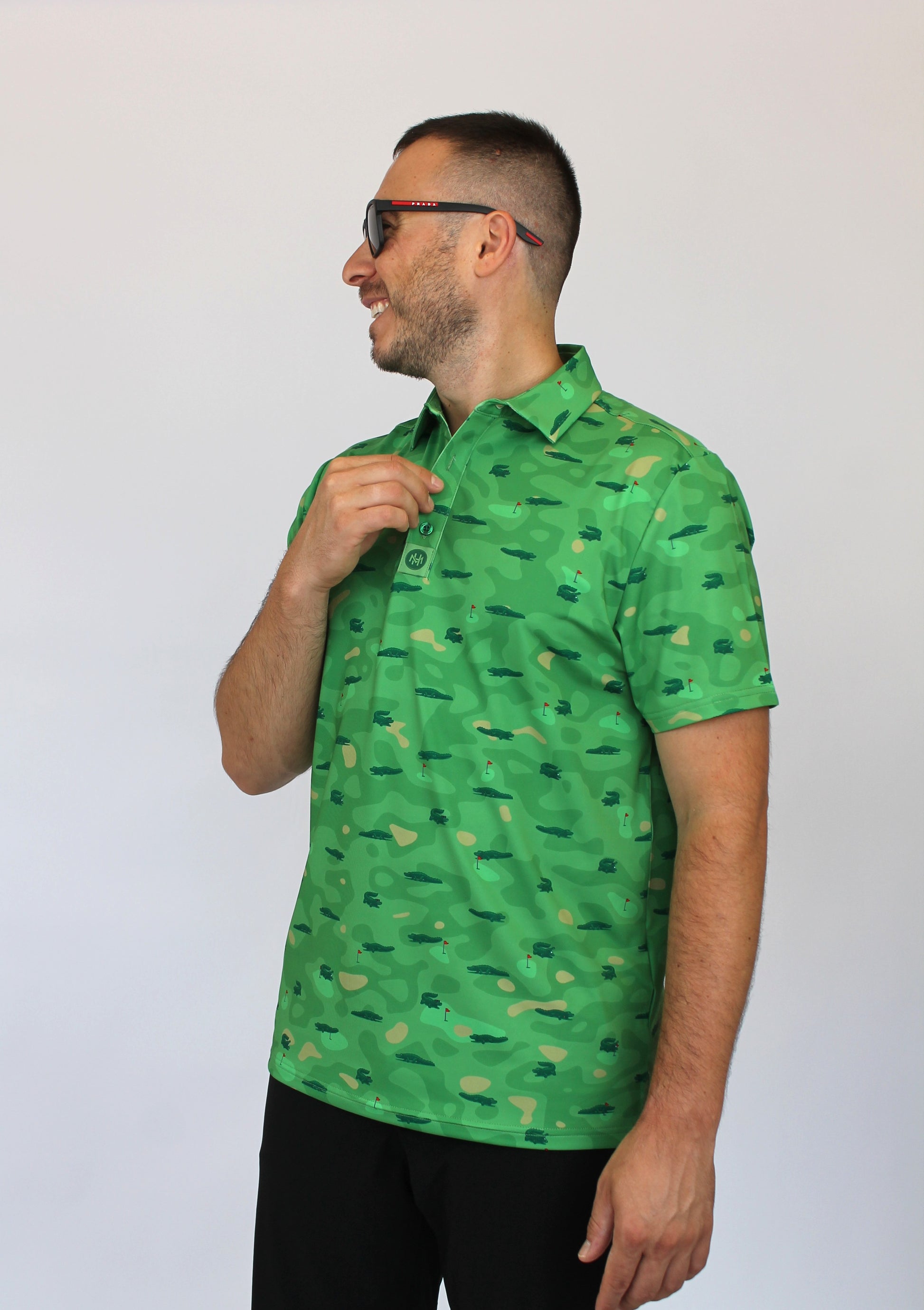 A man wearing a bright camo green short-sleeve button-up shirt with a repeating Alligators on a golf course pattern, paired with black pants and sunglasses, standing against a plain light gray background.