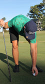 A man on a golf course bends over to place a golf ball on the ground, wearing a green patterned polo shirt, black shorts, and a navy blue cap with white 'Mulligan Hooligan' script.