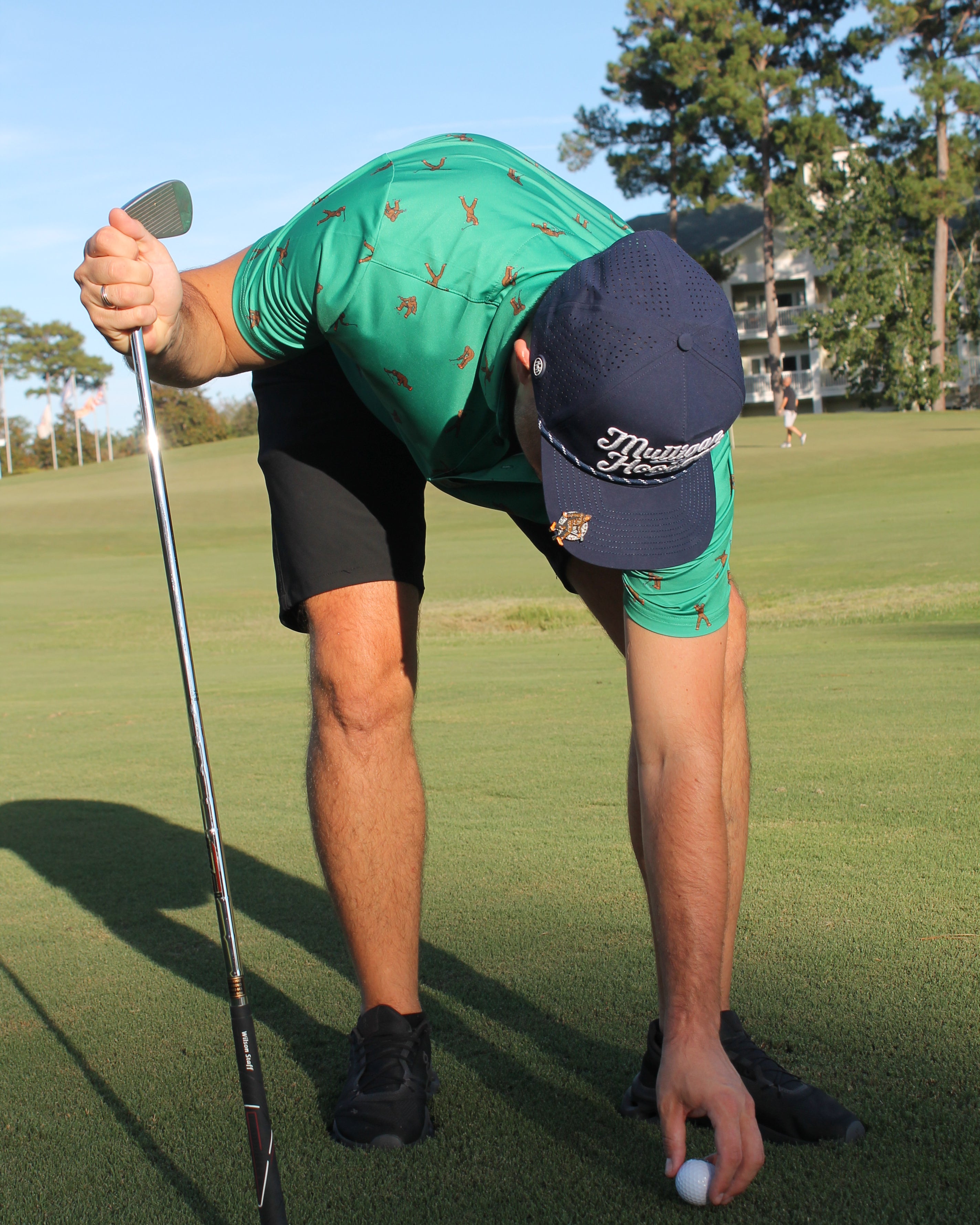 A man on a golf course bends over to place a golf ball on the ground, wearing a green patterned polo shirt, black shorts, and a navy blue cap with white 'Mulligan Hooligan' script.