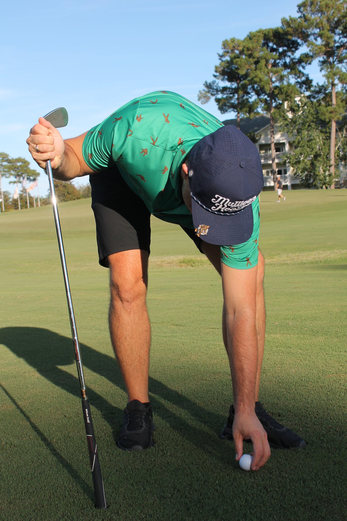 A man on a golf course bends over to place a golf ball on the ground, wearing a green patterned polo shirt, black shorts, and a navy blue cap with white 'Mulligan Hooligan' script.