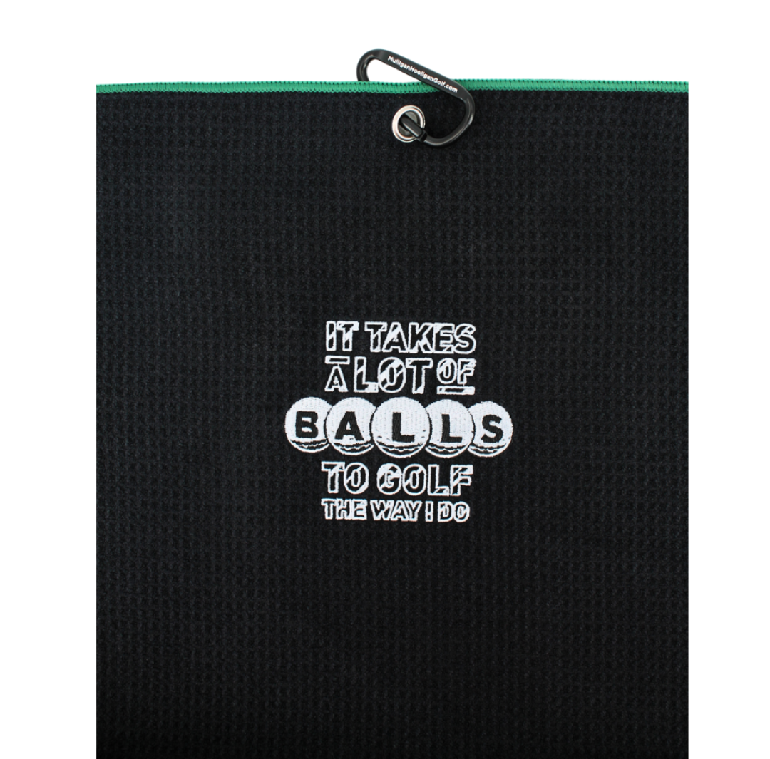 A black textured golf towel with a white embroidered slogan reading 'IT TAKES A LOT OF BALLS TO GOLF THE WAY I DO' and a green zipper trim at the top, shown on a plain white background.