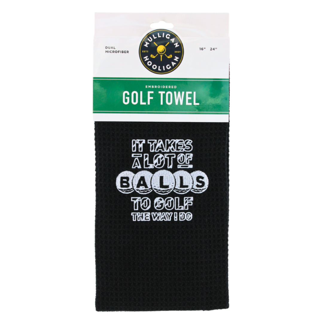 A black waffle-weave golf towel with white printed text reading 'IT TAKES A LOT OF BALLS TO GOLF THE WAY I DO,' hanging from a white and green retail header card. The header card shows the Mulligan Hooligan brand logo and size information.