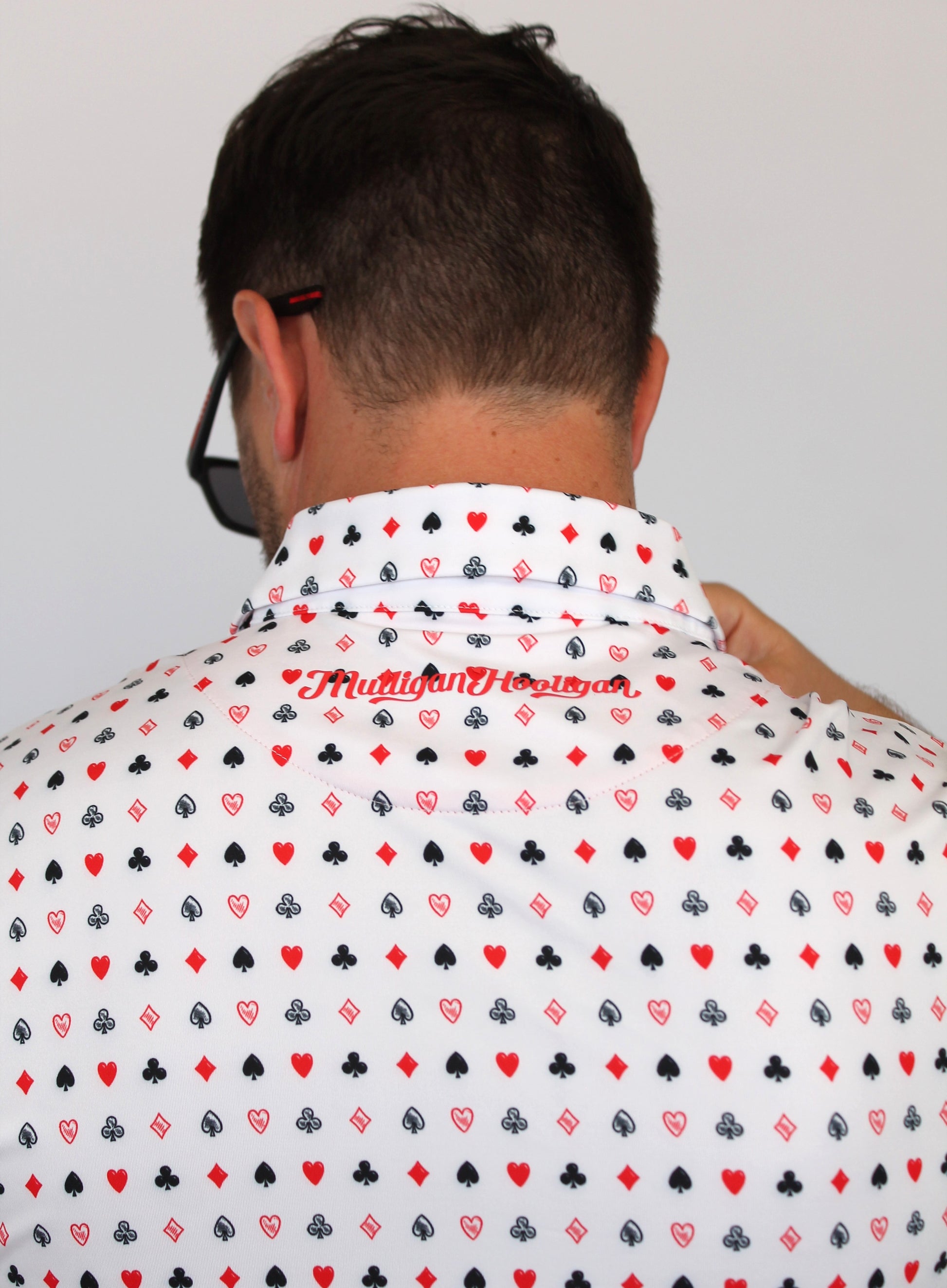 A man viewed from behind wearing a white short-sleeve button-up shirt with an all-over playing card suit pattern in red and black, featuring the brand name 'Mulligan Hooligan' printed in red script across the upper back.