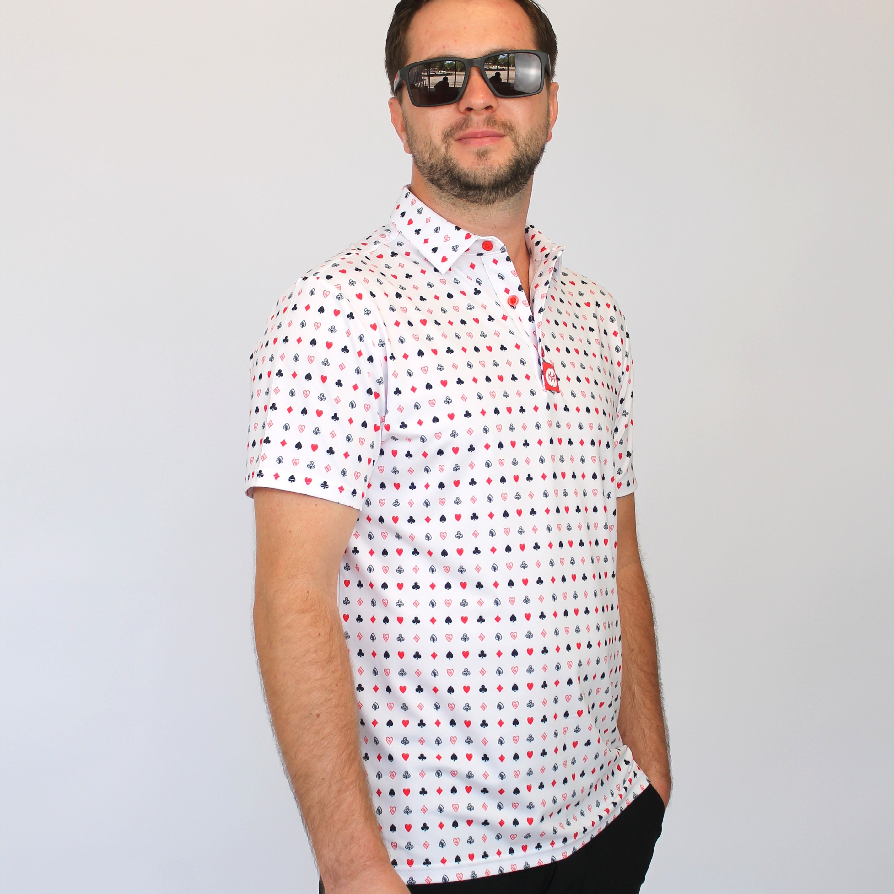 A man wearing a white short-sleeve polo shirt with a small red and blue Playing cards pattern, black pants, and dark sunglasses, standing against a light gray background.
