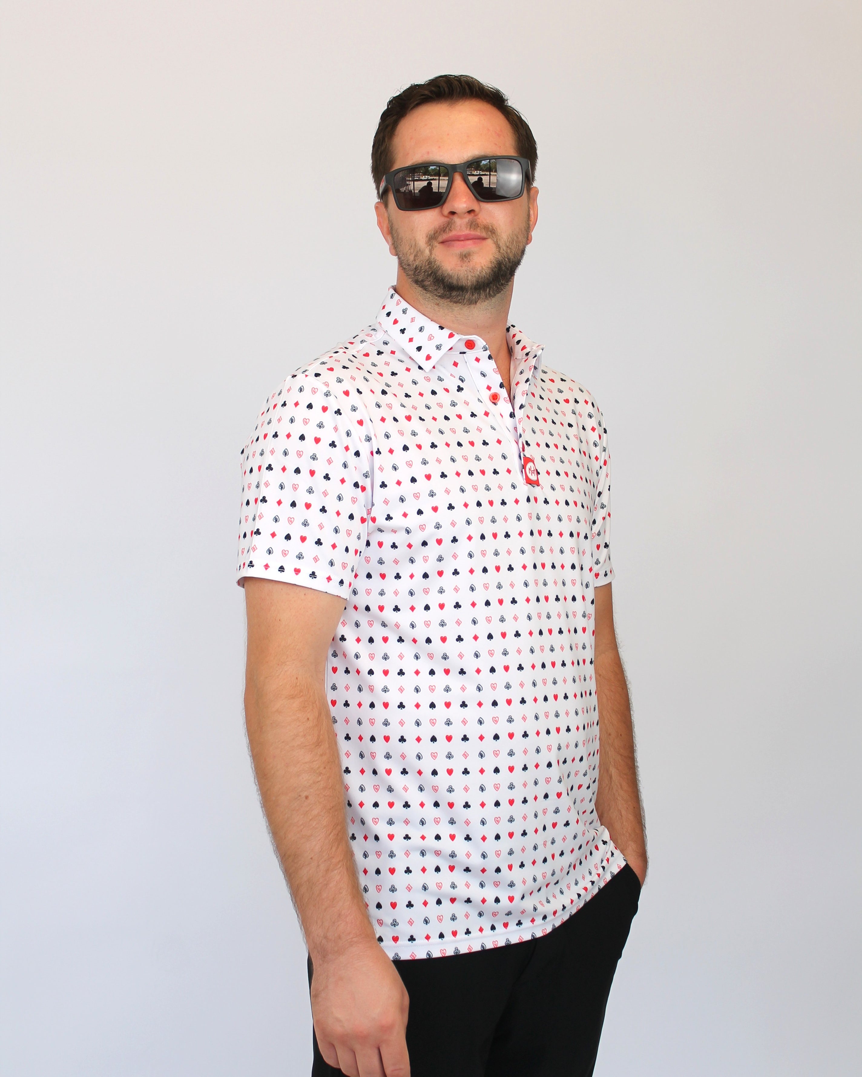 A man wearing a white short-sleeve polo shirt with a small red and blue Playing cards pattern, black pants, and dark sunglasses, standing against a light gray background.