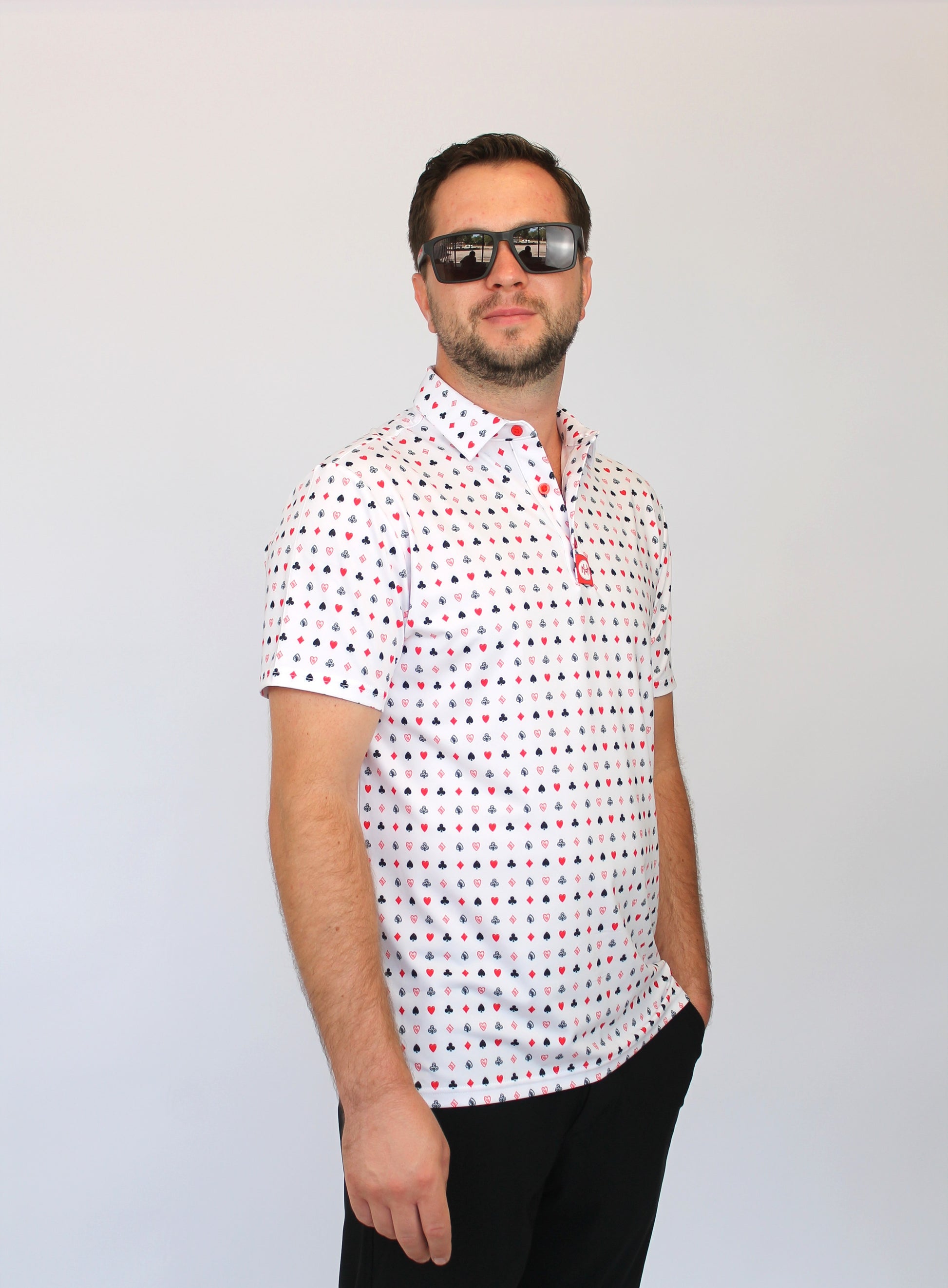 A man wearing a white short-sleeve polo shirt with a small red and blue Playing cards pattern, black pants, and dark sunglasses, standing against a light gray background.