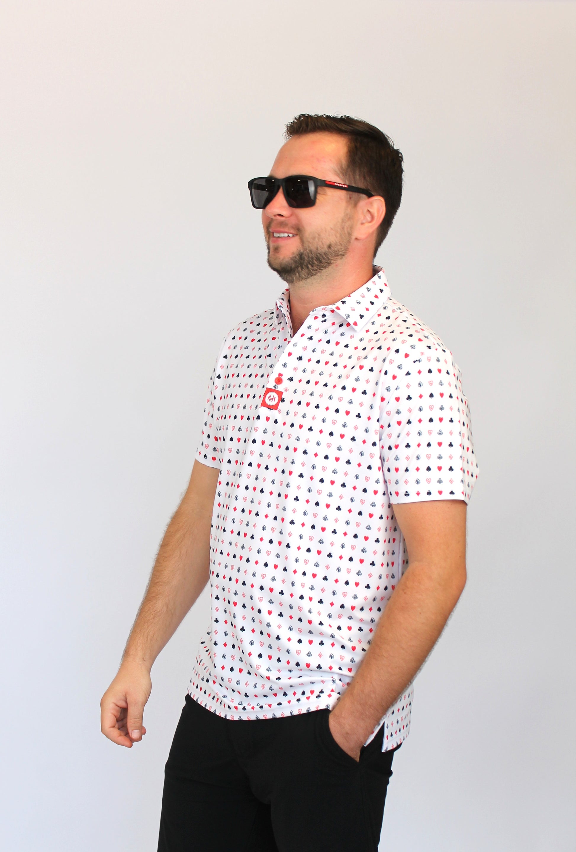 A man wearing a white short-sleeve polo shirt with a red and black playing cards pattern, black pants, and black sunglasses, standing against a plain light gray background.