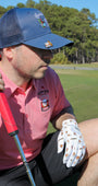 Man in a Mulligan Hooligan 'Sinking Putts' pink polo with a Navy ''Buckets'' cap and a Whiskey Ball Marker, and the Whiskey & Cigars Golf Glove holding a golf club on a golf course