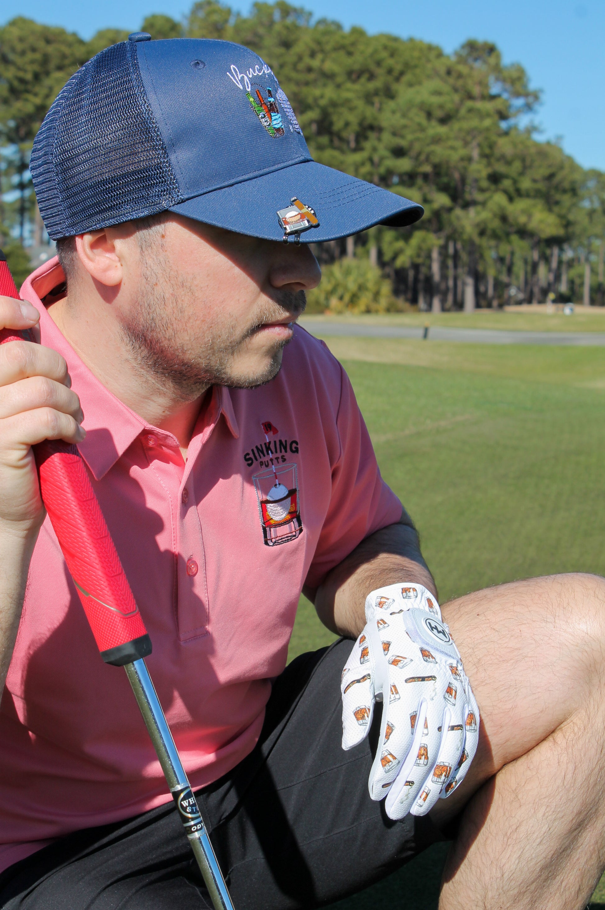 Man in a Mulligan Hooligan 'Sinking Putts' pink polo with a Navy ''Buckets'' cap and a Whiskey Ball Marker, and the Whiskey & Cigars Golf Glove holding a golf club on a golf course