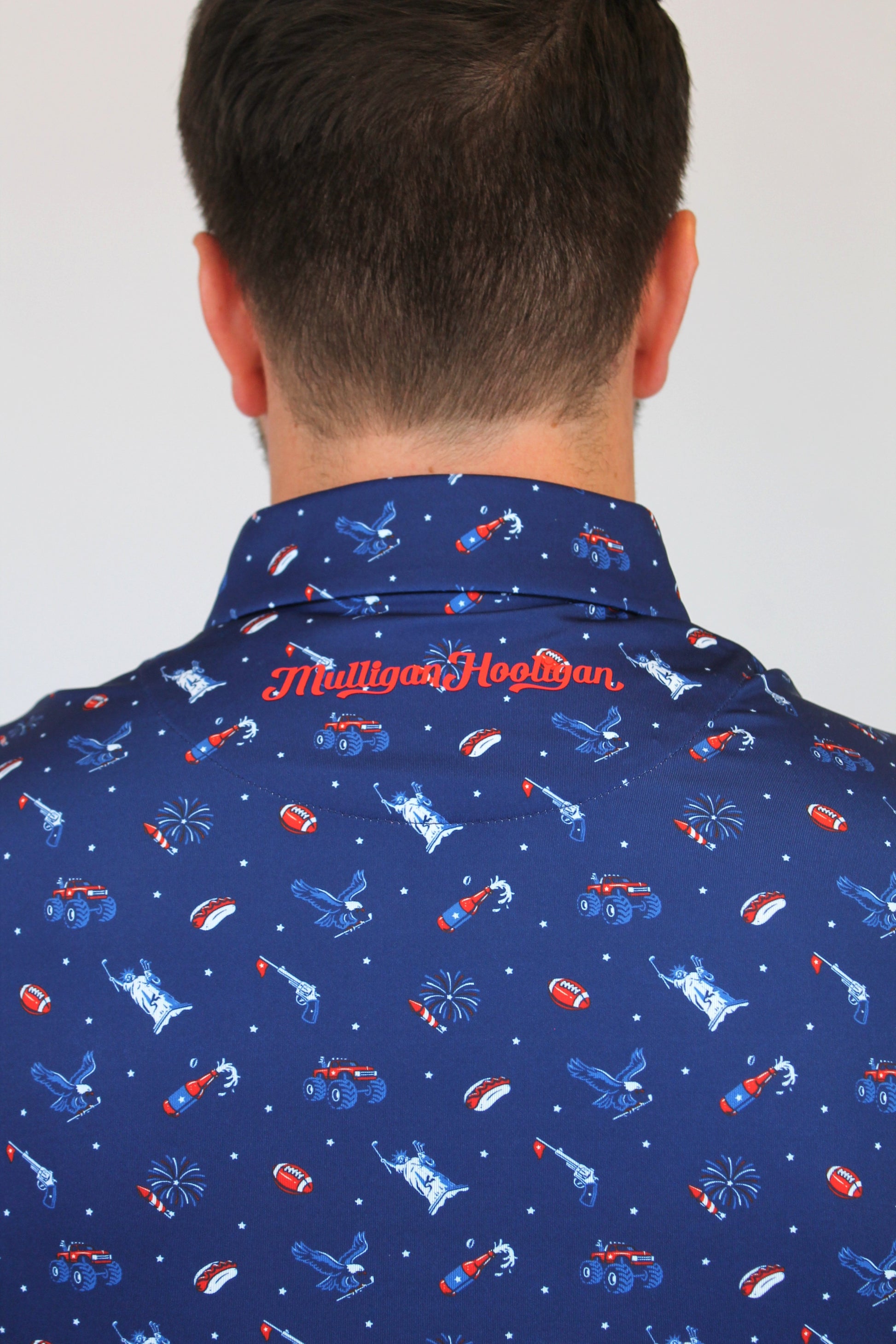 Back view of a man wearing a navy blue collared shirt with a red and white patriotic pattern of rockets, fireworks, and stars. The brand name 'Mulligan Hooligan' is embroidered in red on the back of the collar.
