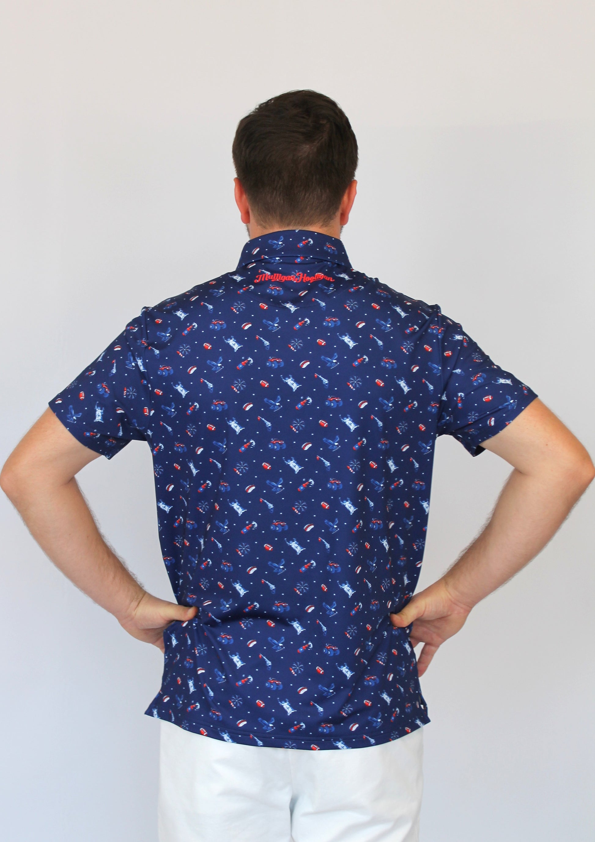 A man viewed from behind wearing a navy blue short-sleeve button-up shirt with a small repeating red and white patriotic-theme pattern, paired with white pants, against a plain light gray background.