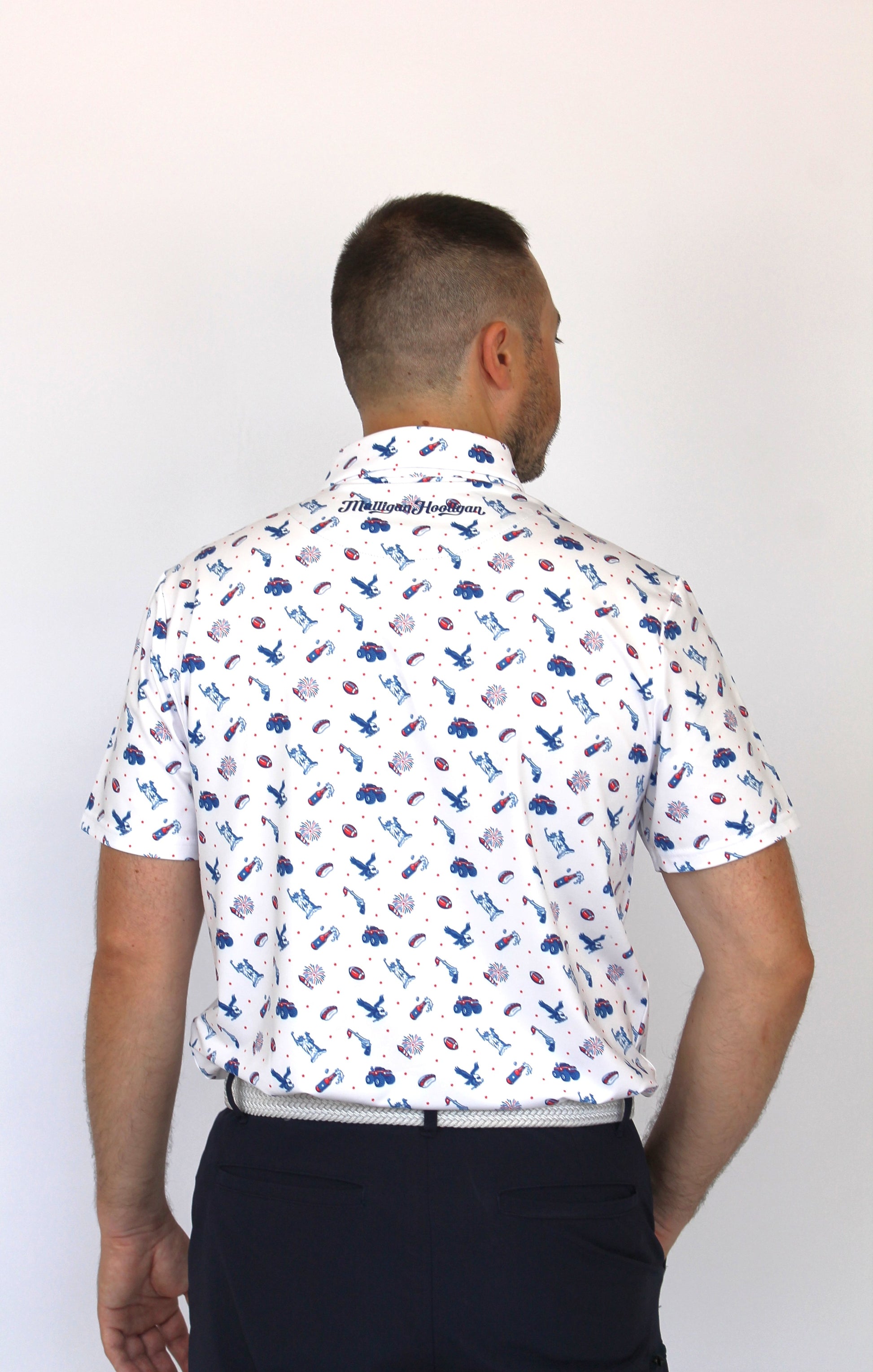 A man viewed from behind wearing a white short-sleeve collared shirt with a patriotic-theme all-over print of small blue, red, and gray icons, paired with dark navy pants, against a plain white background.