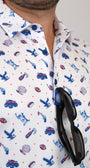 A close-up of a man wearing a white short-sleeve button-up shirt with a blue and red patriotic-themed print of cars, fireworks, and eagles. Black sunglasses are tucked into the collar.