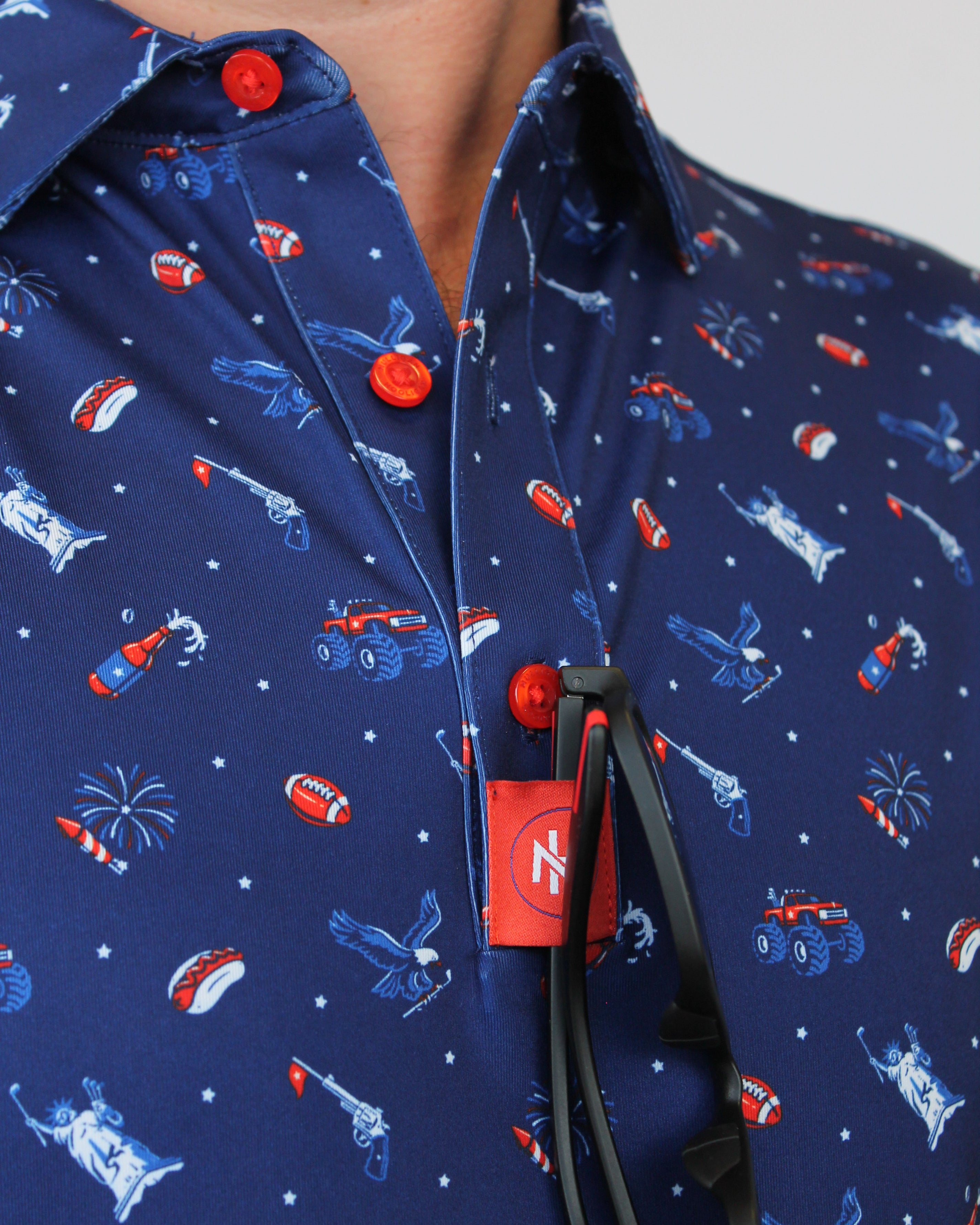 Close-up of a navy blue shirt with a patriotic red, white, and blue pattern of sports figures, stars, and flags, featuring red snap buttons and a red 'MH' brand tag.