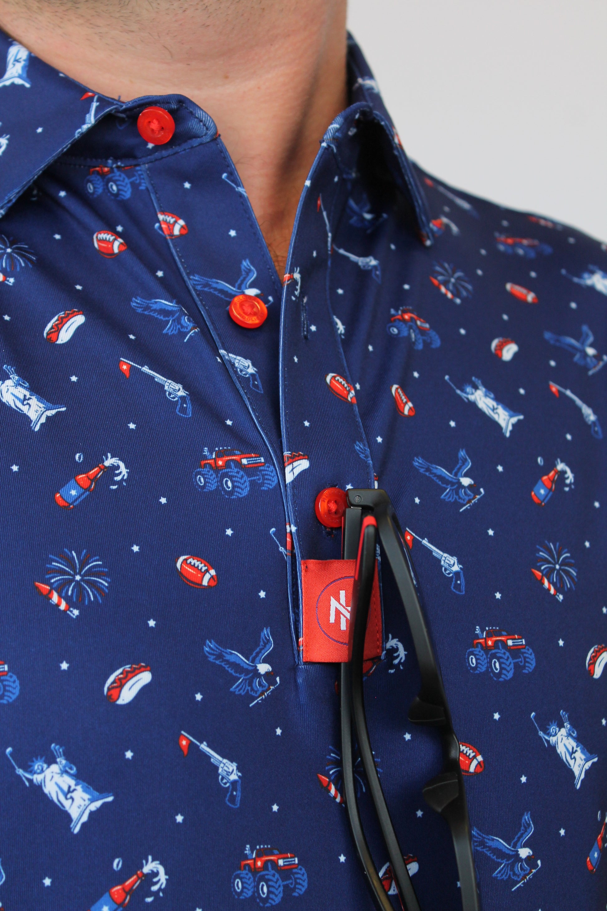 Close-up of a navy blue shirt with a patriotic red, white, and blue pattern of sports figures, stars, and flags, featuring red snap buttons and a red 'MH' brand tag.