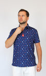 A man wearing a navy blue short-sleeve button-up shirt with a repeating red and white patriotic-theme / USA pattern, paired with white pants, standing against a plain light gray background.