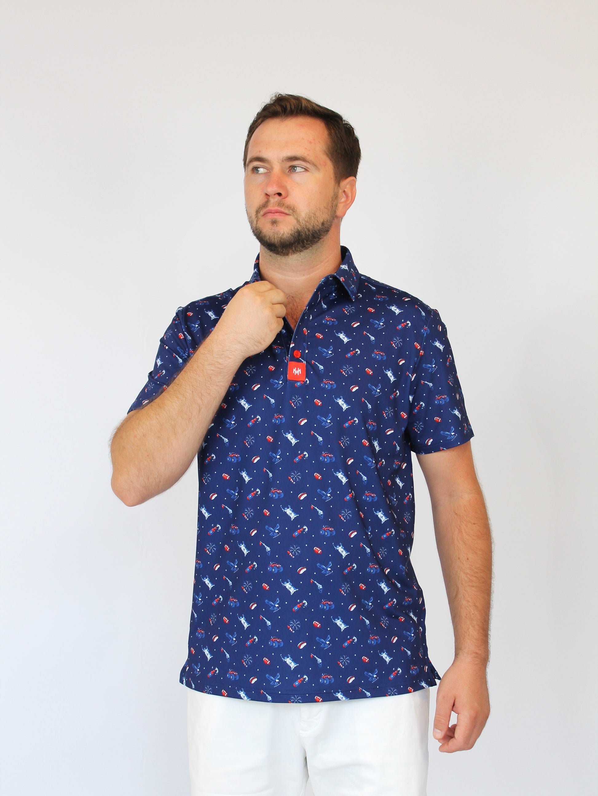 A man wearing a navy blue short-sleeve button-up shirt with a repeating red and white patriotic-theme / USA pattern, paired with white pants, standing against a plain light gray background.