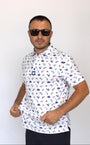 A man wearing a white short-sleeve button-up shirt with a blue and red patriotic-theme print of fireworks, hot-dogs, golfing liberty statue,  beer bottle and eagles, paired with dark sunglasses and dark pants, standing against a plain white background.