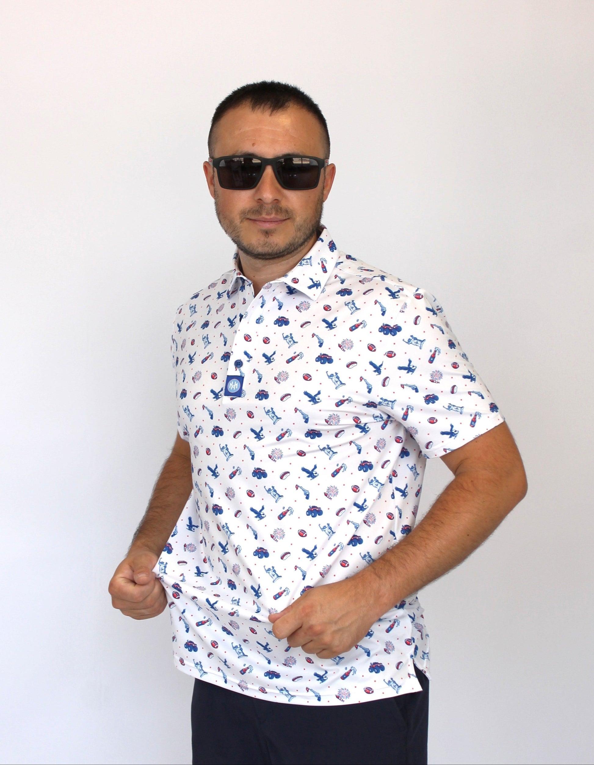 A man wearing a white short-sleeve button-up shirt with a blue and red patriotic-theme print of fireworks, hot-dogs, golfing liberty statue,  beer bottle and eagles, paired with dark sunglasses and dark pants, standing against a plain white background.