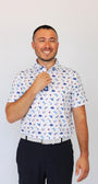 A man wearing a white short-sleeve button-up shirt with a blue and red USA pattern, paired with dark trousers, standing against a plain light gray background.

