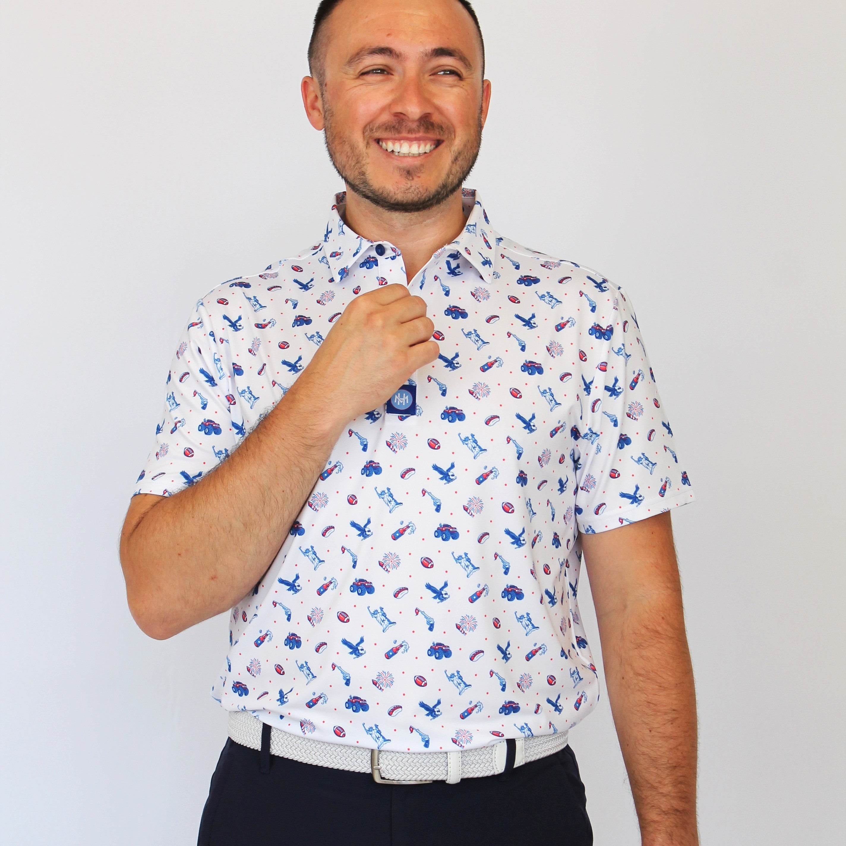 A man wearing a white short-sleeve button-up shirt with a blue and red USA pattern, paired with dark trousers, standing against a plain light gray background.
