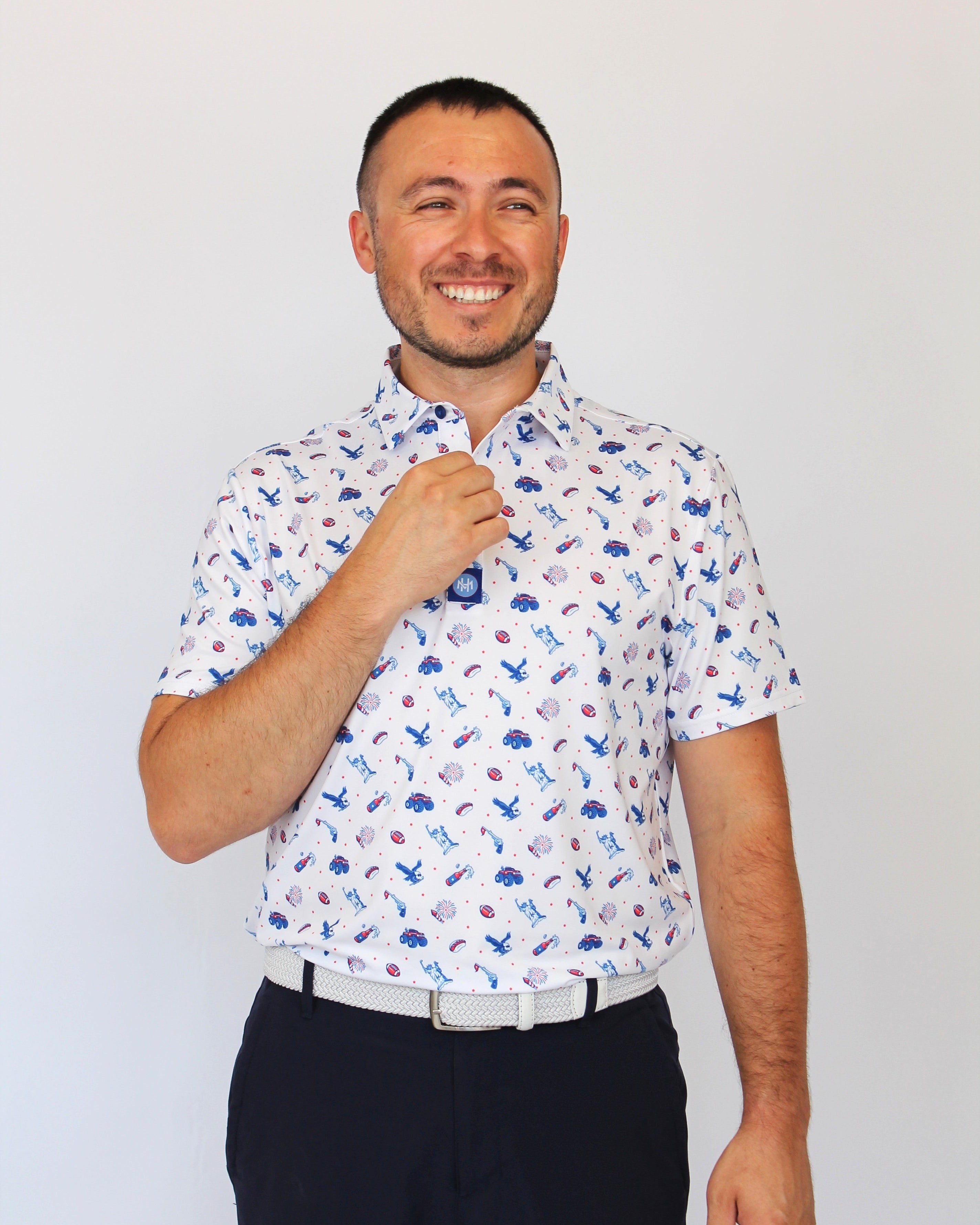 A man wearing a white short-sleeve button-up shirt with a blue and red USA pattern, paired with dark trousers, standing against a plain light gray background.

