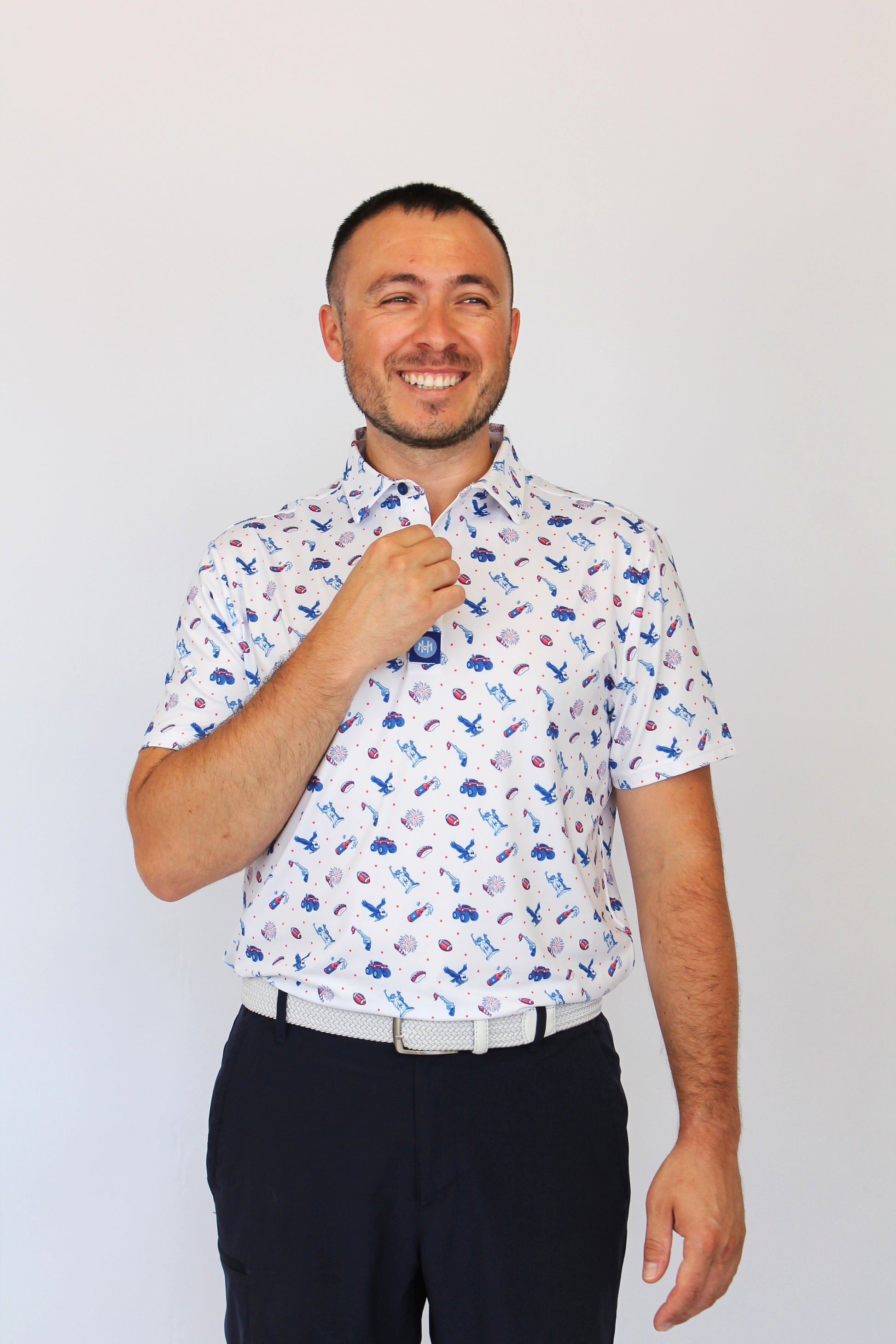 A man wearing a white short-sleeve button-up shirt with a blue and red USA pattern, paired with dark trousers, standing against a plain light gray background.
