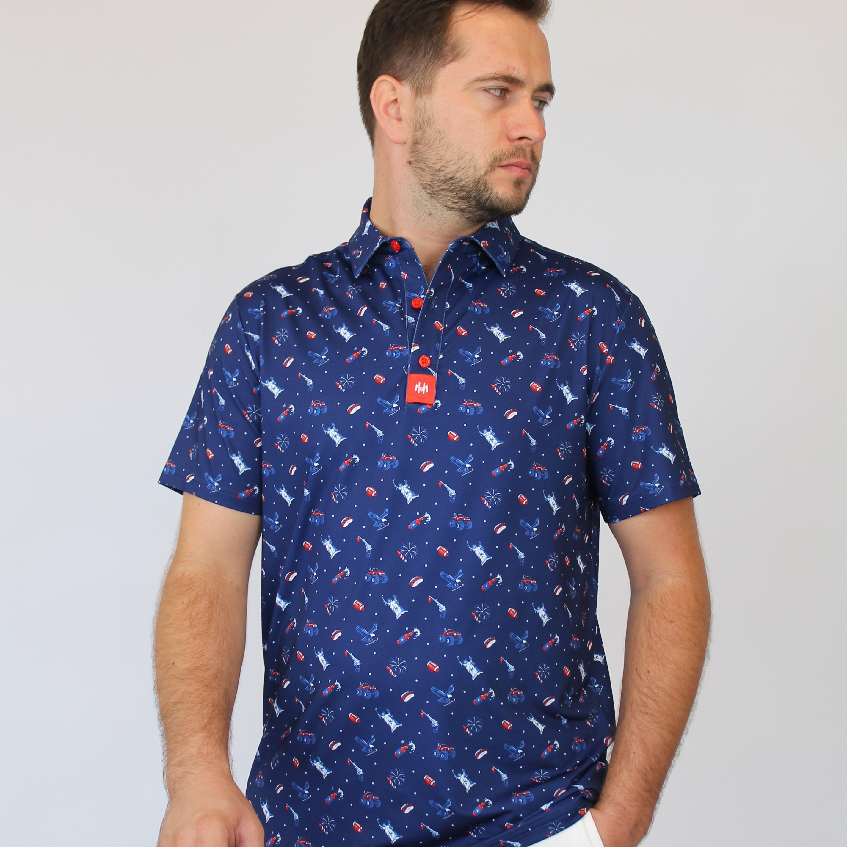 A man wearing a short-sleeve button-up shirt with a blue, red, and white patriotic-theme pattern, paired with white trousers, standing against a light gray background.