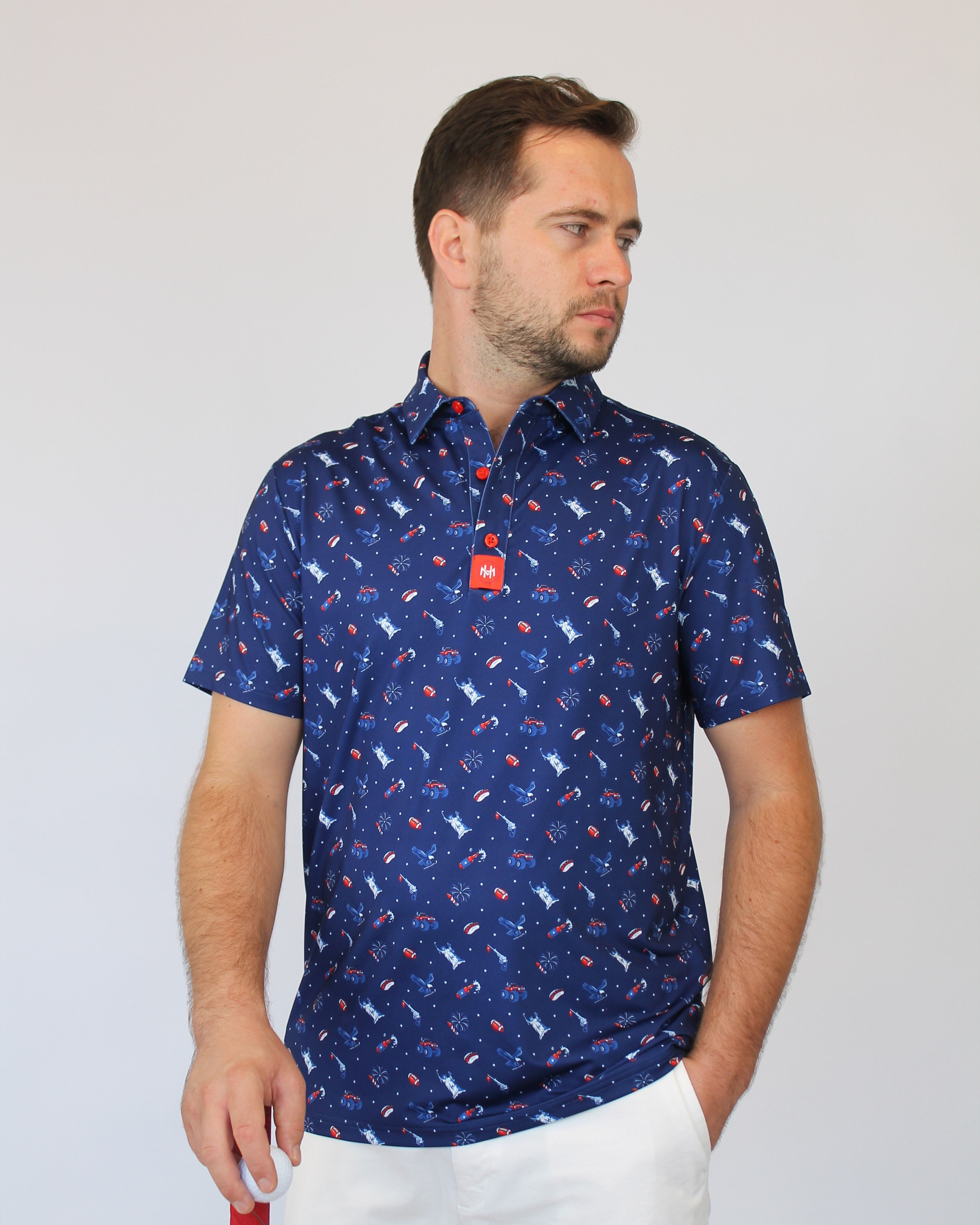 A man wearing a short-sleeve button-up shirt with a blue, red, and white patriotic-theme pattern, paired with white trousers, standing against a light gray background.