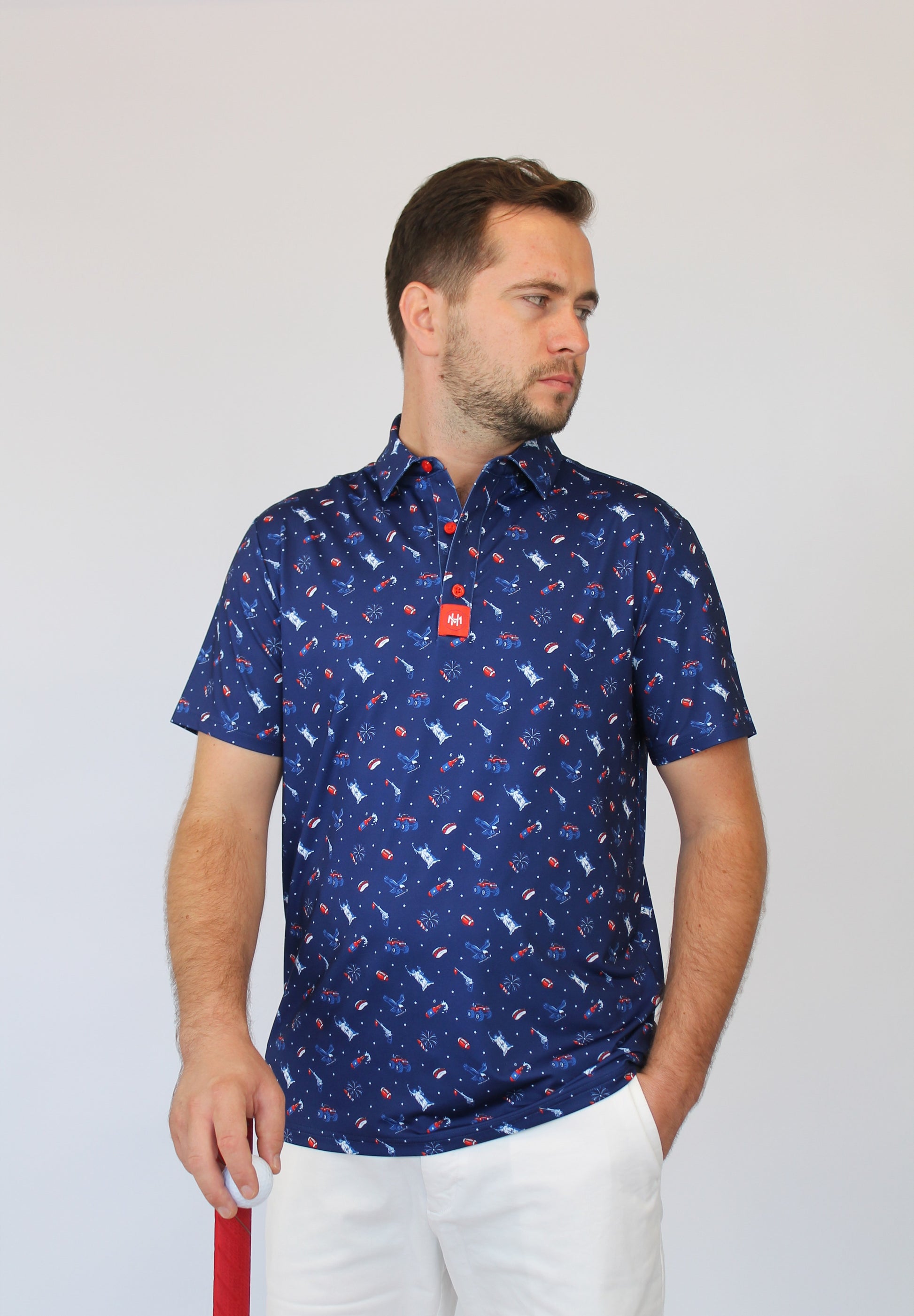 A man wearing a short-sleeve button-up shirt with a blue, red, and white patriotic-theme pattern, paired with white trousers, standing against a light gray background.