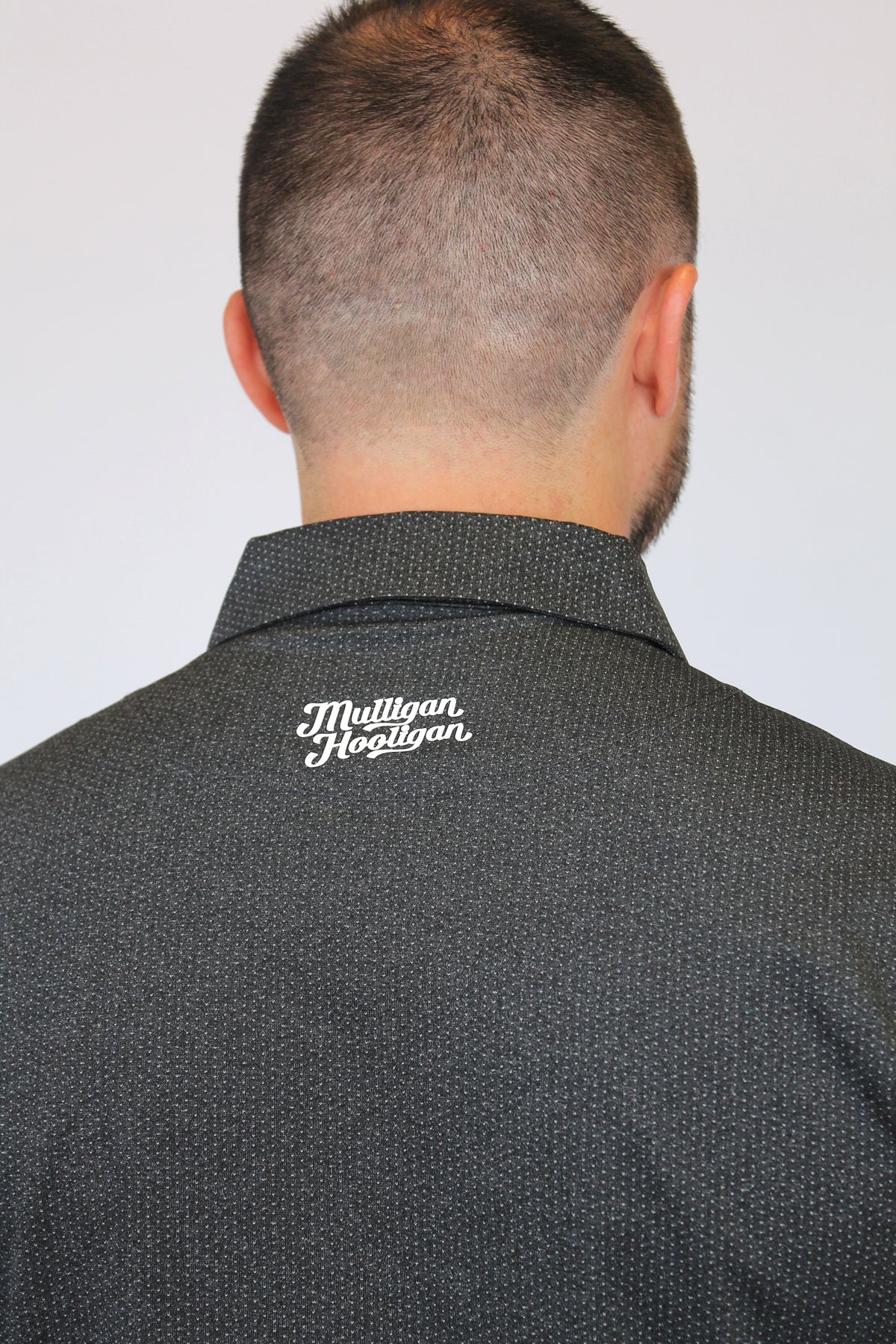 Back view of a man wearing a dark gray textured collared shirt with 'Mulligan Hooligan' embroidered in white script on the upper back, against a plain light gray background.