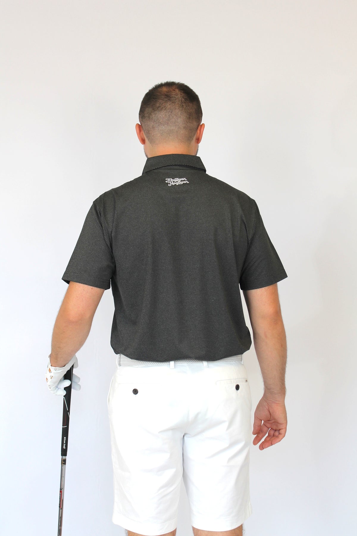 A man viewed from behind wearing a dark charcoal gray short-sleeve polo shirt and white shorts, holding a golf club against a plain white background.