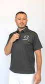 A man wearing a dark gray short-sleeve polo shirt with a white embroidered logo on the left chest, paired with white pants, standing against a plain white background.