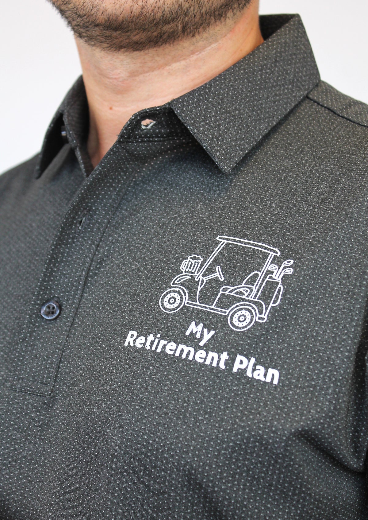 A close-up of a man wearing a dark gray micro-patterned button-down shirt with a white printed golf cart and the text 'My Retirement Plan' on the chest.