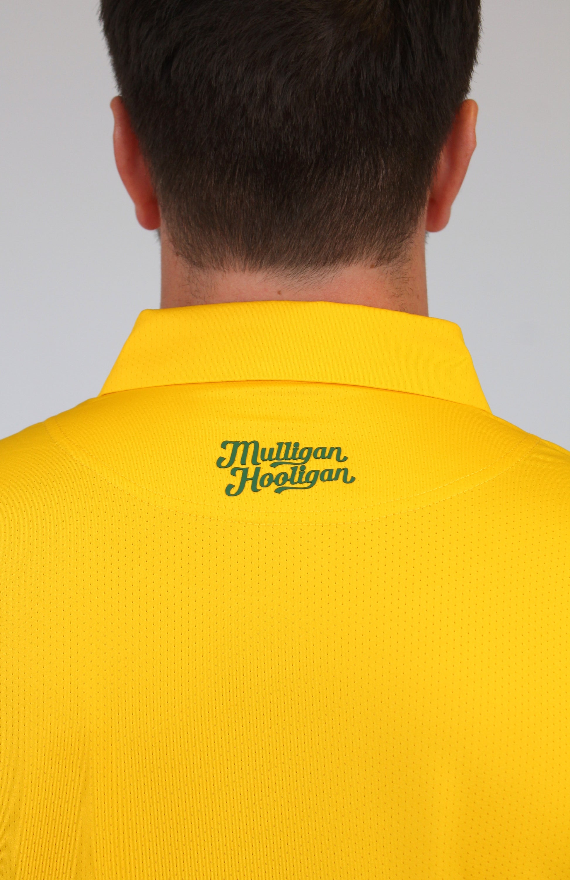 Back view of a person wearing a bright yellow collared shirt with 'Mulligan Hooligan' embroidered in green script on the upper back, against a light gray background.