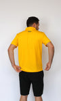 A man viewed from behind wearing a bright yellow short-sleeve polo shirt and black shorts, standing against a plain white wall.