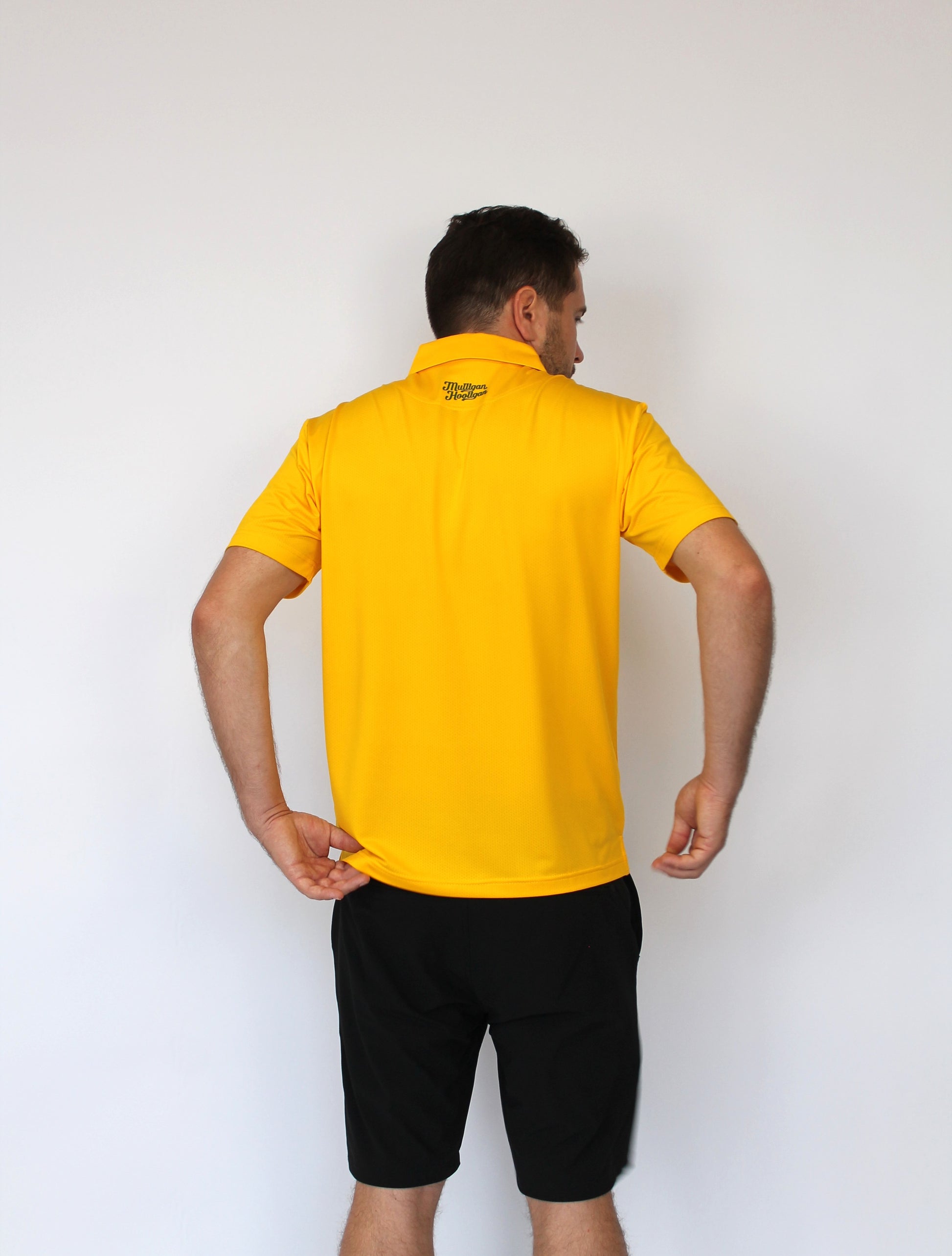 A man viewed from behind wearing a bright yellow short-sleeve polo shirt and black shorts, standing against a plain white wall.