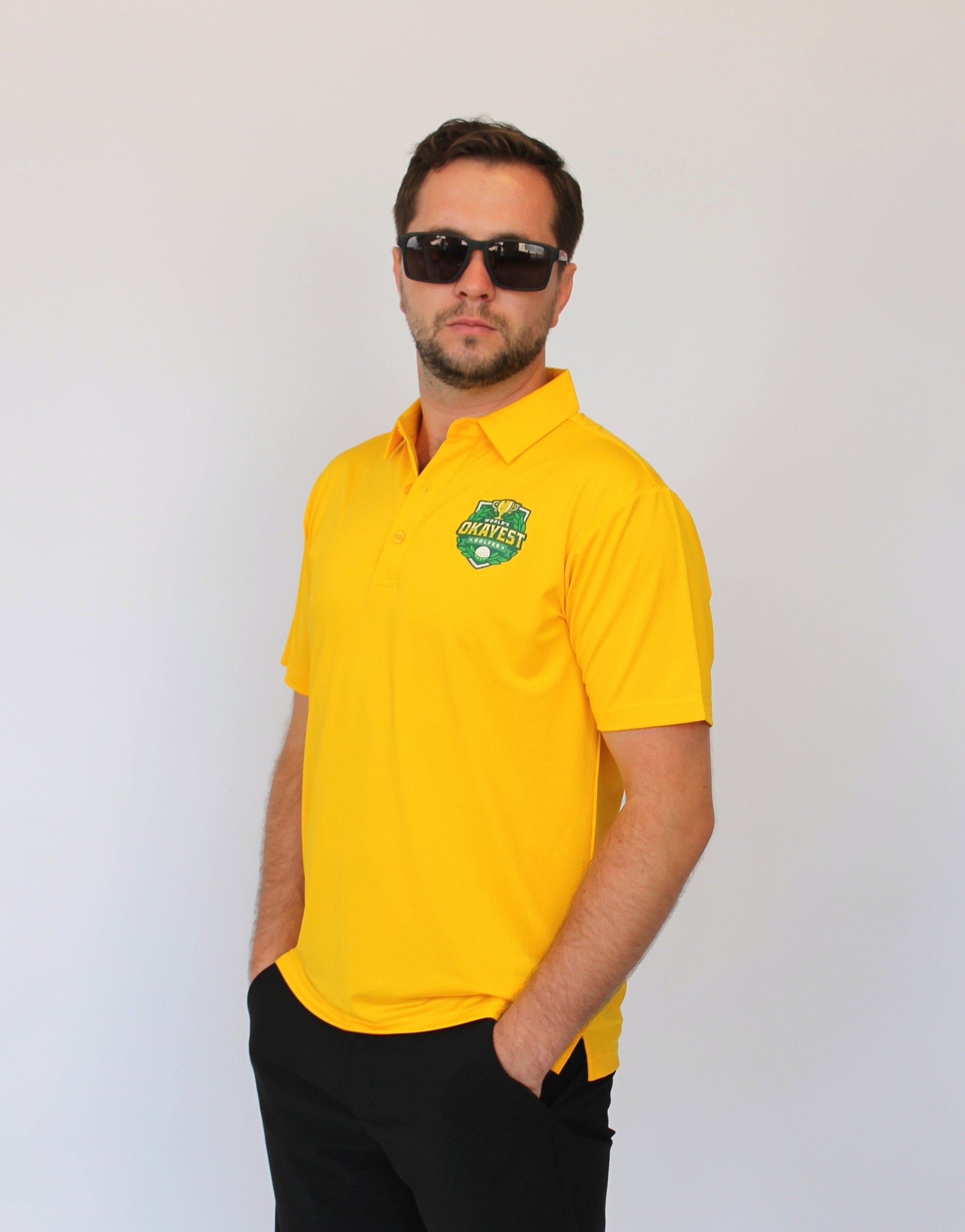 A man wearing a bright yellow short-sleeve polo shirt with a green and white 'Okayest Golfer' Graphic Golf Polo crest on the left chest, paired with black shorts and sunglasses, standing against a plain white background.