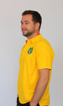 A man wearing a bright yellow short-sleeve polo shirt with a green and white shield logo on the left chest, standing against a plain light gray background.