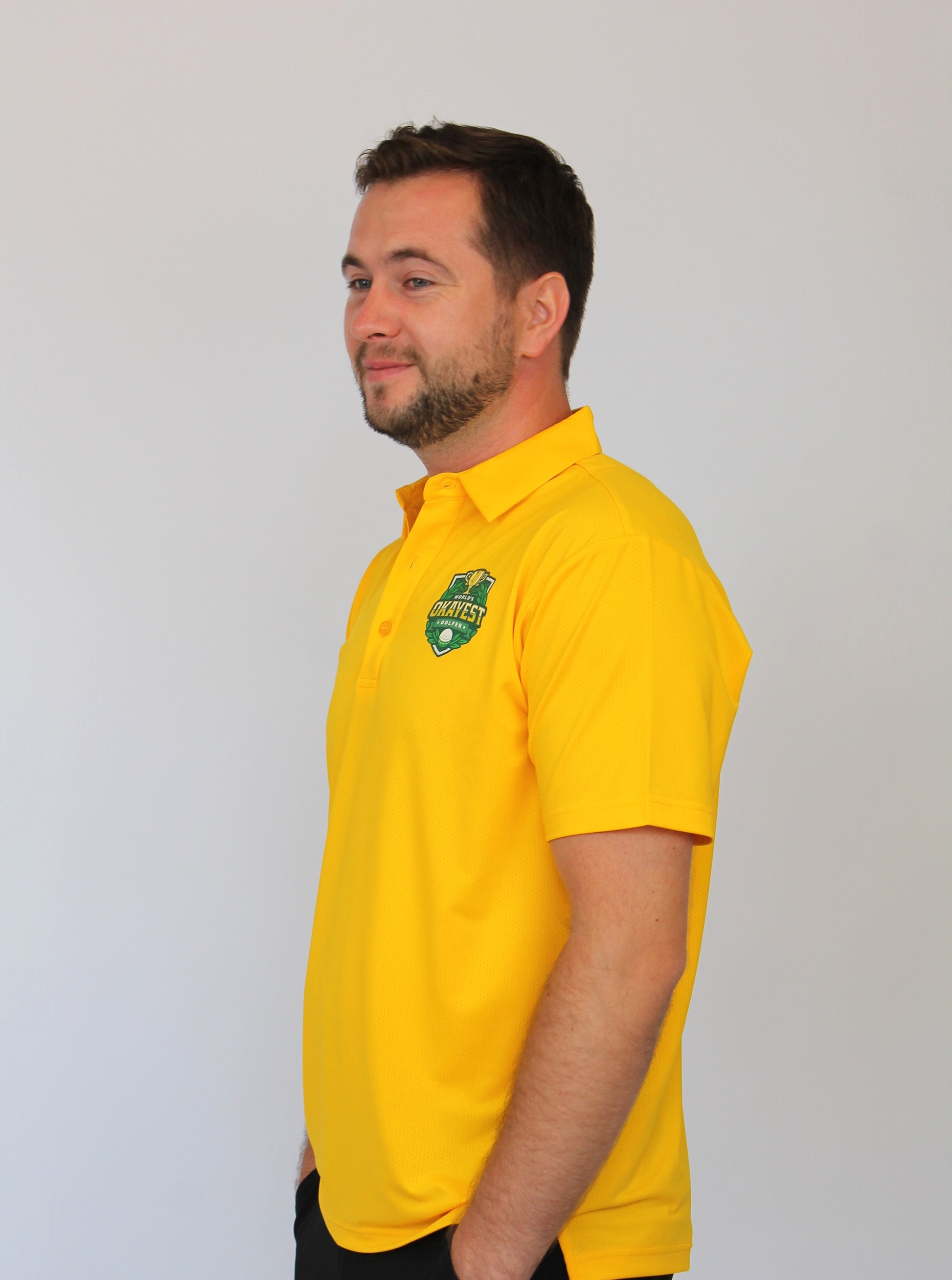 A man wearing a bright yellow short-sleeve polo shirt with a green and white shield logo on the left chest, standing against a plain light gray background.