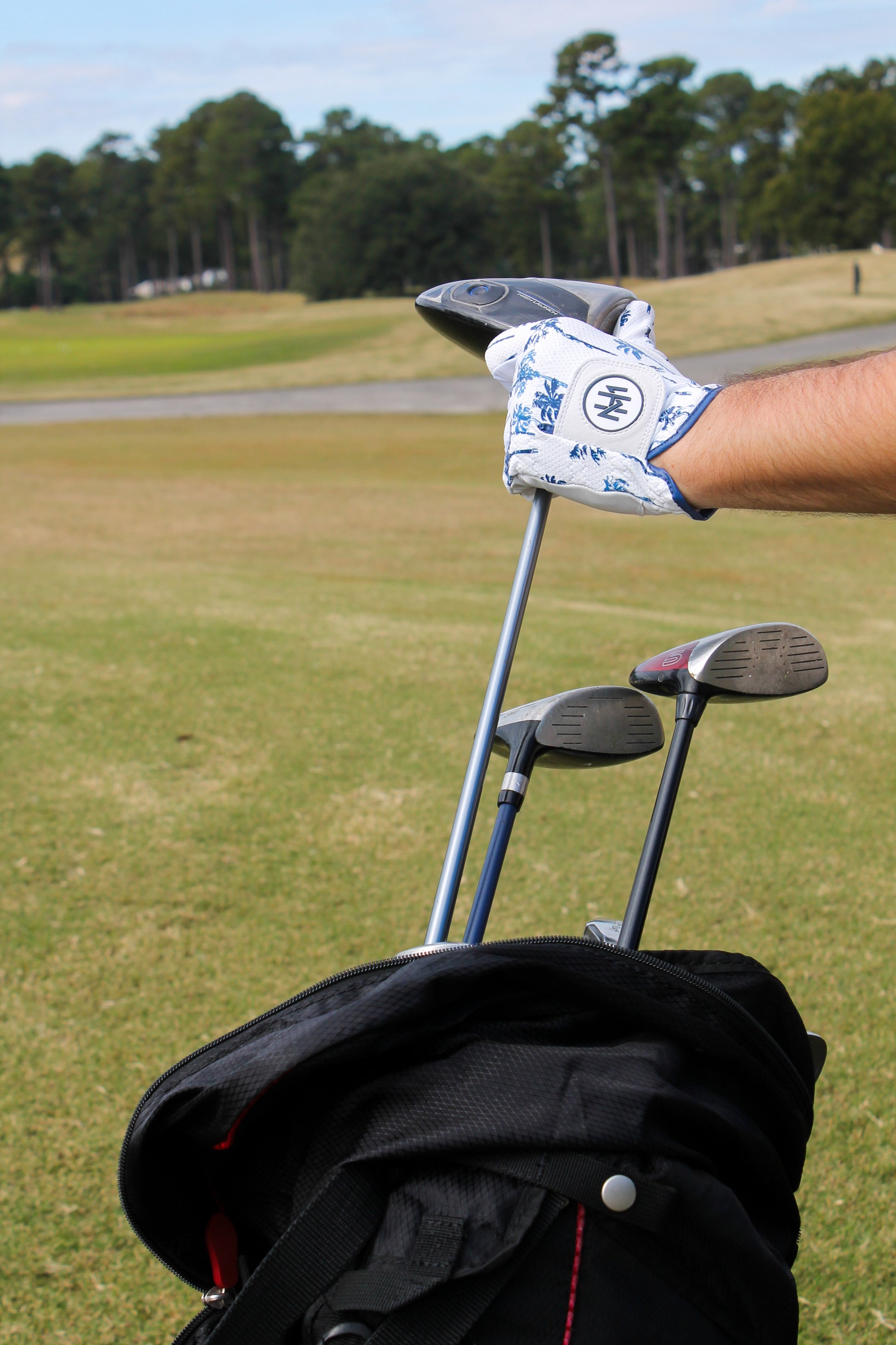 A hand in a Mulligan Hooligan "Palm Par" golf glove pulling a golf club out of a bag on a golf course.