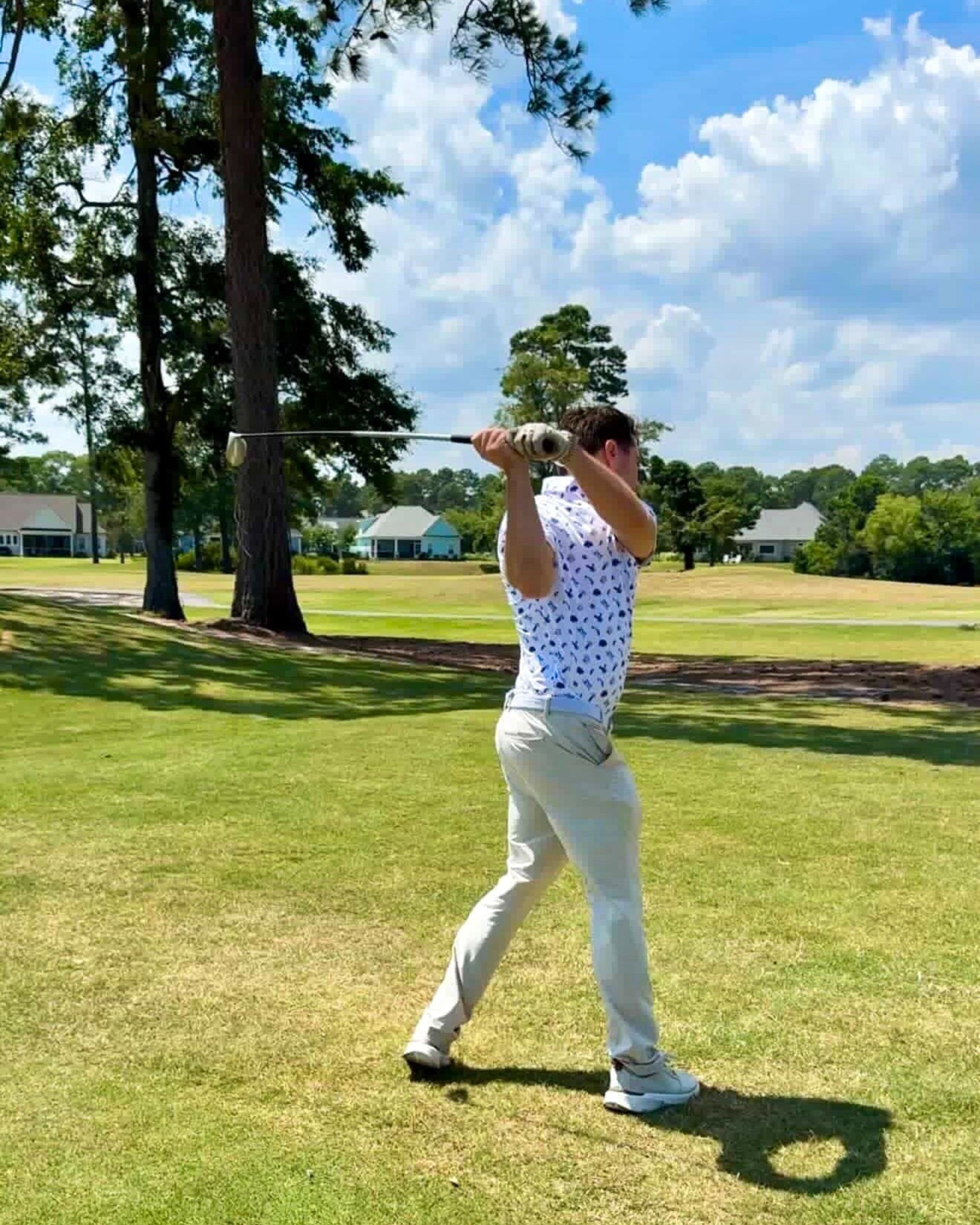 A man wearing a white short-sleeve polo shirt with a blue pattern and beige trousers is captured mid-swing on a golf course under a blue sky with clouds.