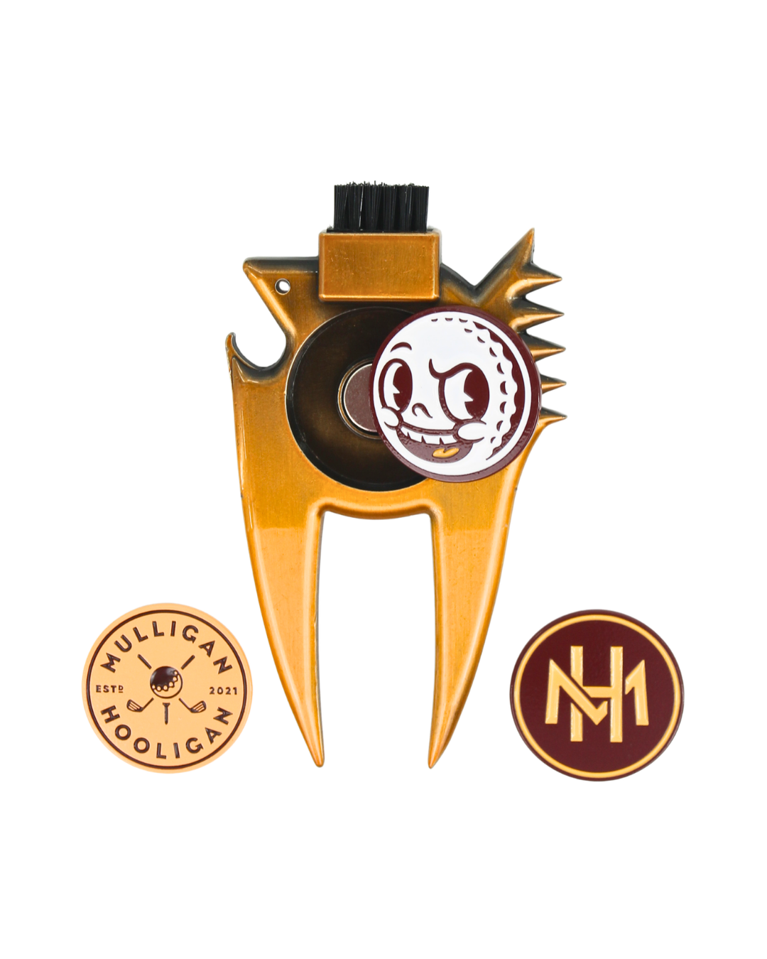 Gold metal golf divot tool with a black bristle brush on top, a circular white and maroon golf ball emblem in the center, and two circular gold and maroon logo patches below on a white background.