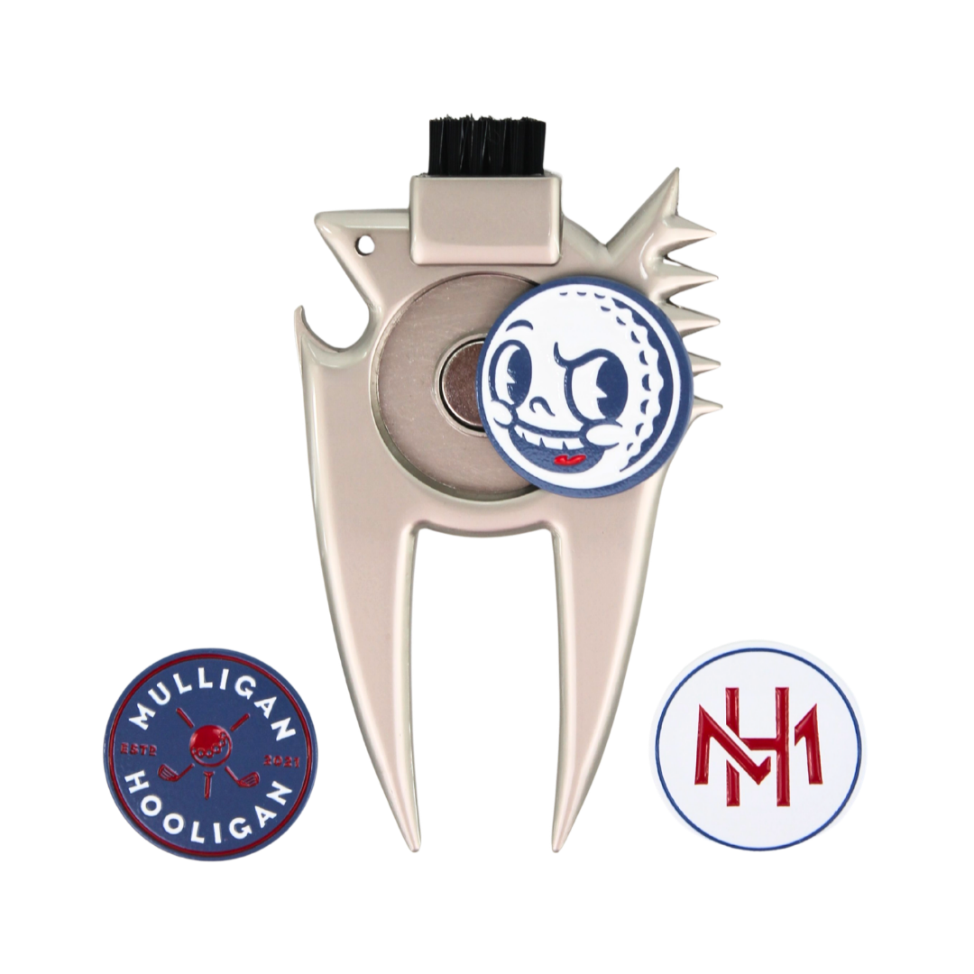A silver metal golf divot tool with a black bristle brush on top, featuring a circular golf ball graphic with a cartoon face on the center. Two circular decals are shown below: one blue and red reading 'MULLIGAN HOOLIGAN' and one white and red with 'MH'.