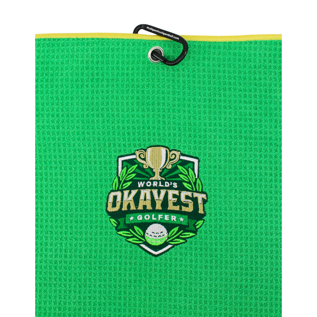A bright green textured golf towel with a black hanging loop and a yellow edge trim, featuring a central embroidered crest that reads 'World's Okayest Golfer' with a trophy, golf ball, and green leaves.