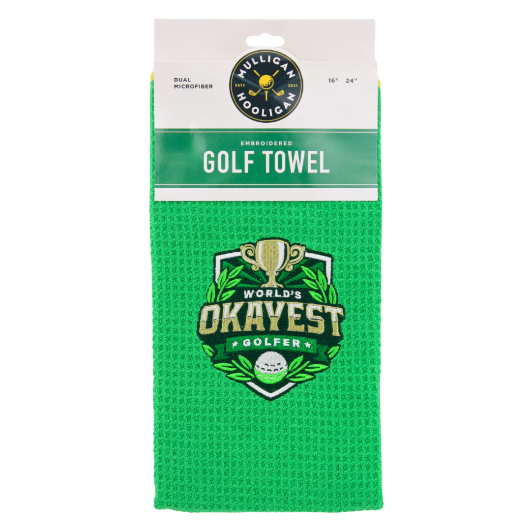 A bright green waffle-weave golf towel in its retail packaging, featuring a 'World's Okayest Golfer' logo on the front.